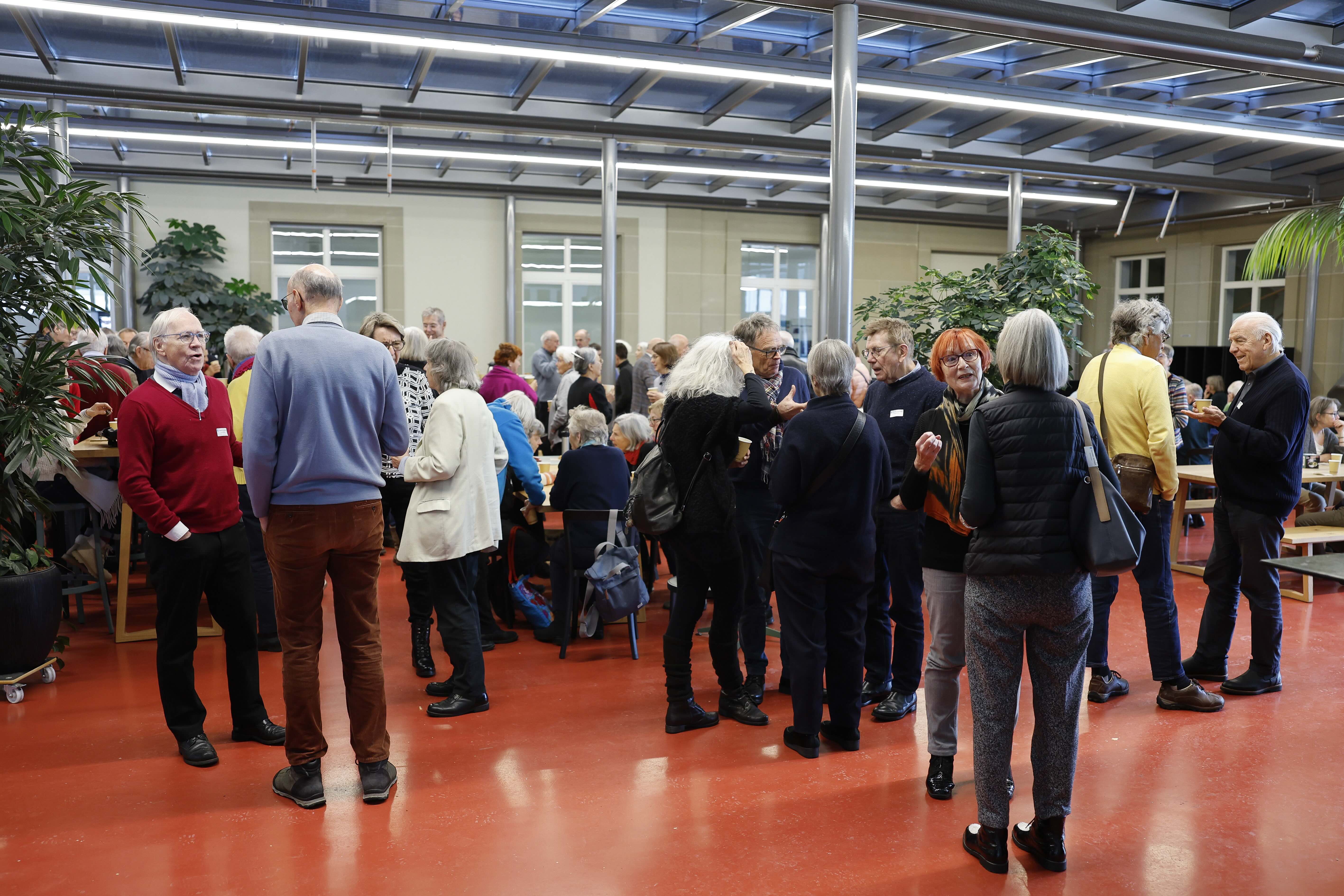 Verschiedene Menschen stehen in einem Foyer mit rotem Boden und sind ins Gespräch vertieft. Einige grüne Zimmerpflanzen sind aufgestellt.
