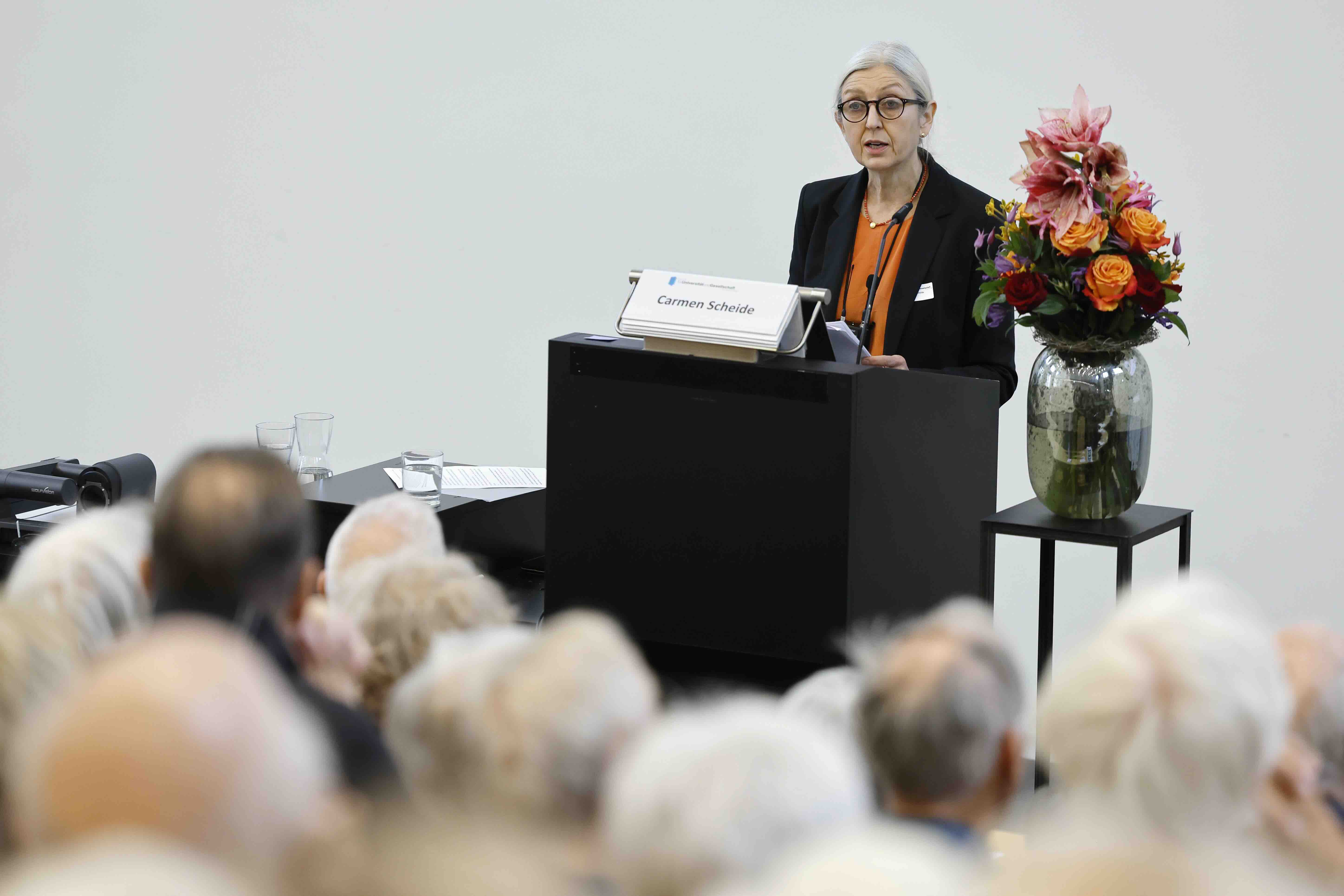 Eine Frau mit grauen Haaren und Brille spricht an einem Rednerpult vor einem Publikum. Neben ihr steht ein großer Blumenstrauß mit orangefarbenen und rosa Blüten. Die Szene zeigt einen Vortrag in einem hellen Raum, im Vordergrund sind unscharf die Zuhörenden zu sehen.