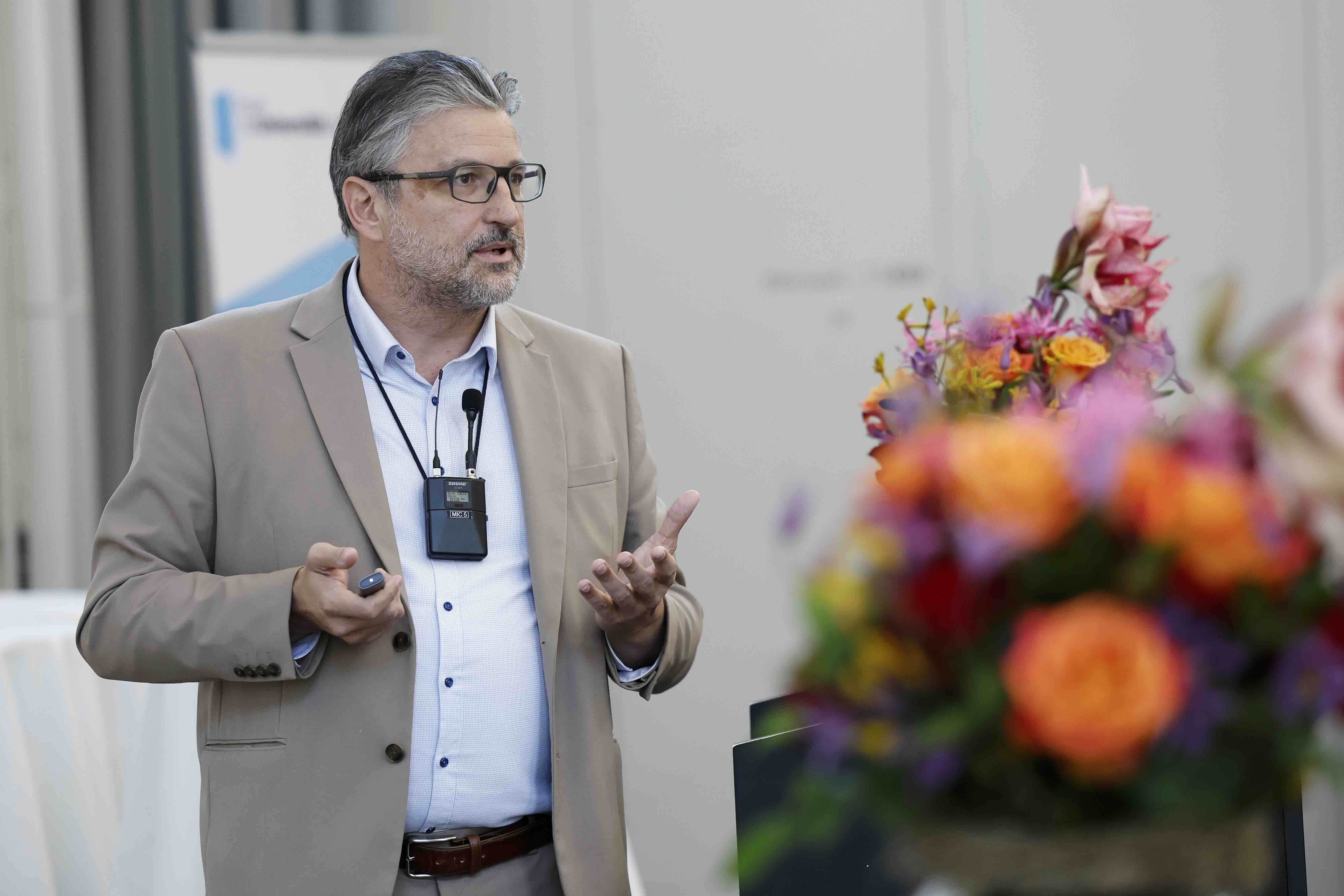 Ein Mann mit Brille und grauem Haar steht bei einem Vortrag und gestikuliert mit den Händen. Er trägt ein beige­farbenes Sakko und ein Headset-Mikrofon. Rechts im Bild ist unscharf ein bunter Blumenstrauß zu sehen, der den Vordergrund dominiert. Der Hintergrund ist hell und schlicht.