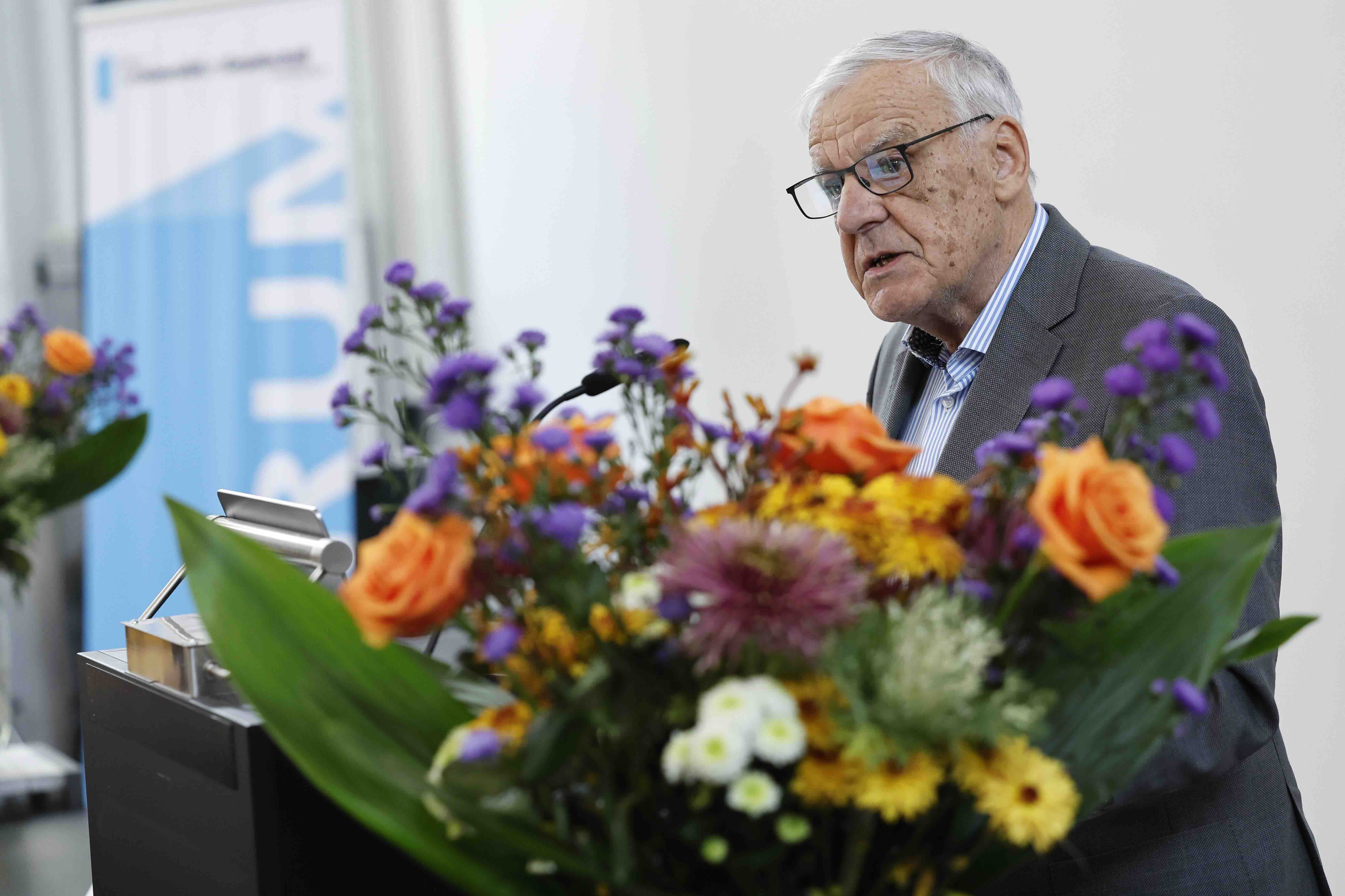 Ein älterer Mann mit grauen Haaren und Brille spricht an einem Rednerpult. Er trägt einen Anzug und wirkt konzentriert. Im Vordergrund steht ein großer Blumenstrauß mit orangen Rosen, gelben und violetten Blüten. Im Hintergrund ist ein Banner mit der Aufschrift „Forum“ zu sehen.