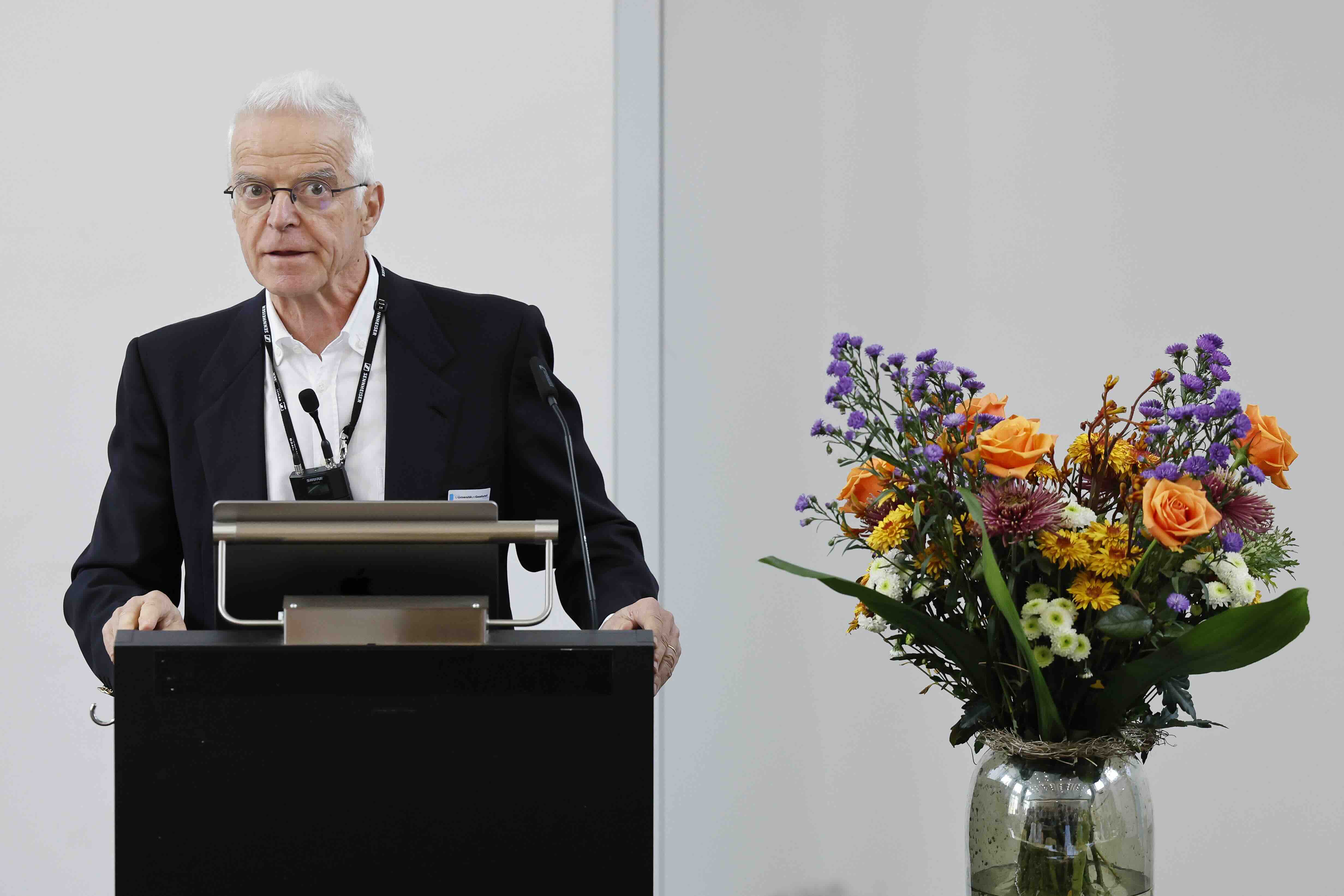 Ein älterer Mann mit grauen Haaren und Brille steht an einem Rednerpult und spricht vor Publikum. Er trägt einen dunklen Anzug und ein weisses Hemd. Rechts neben ihm steht ein grosser Blumenstrauss mit orangen Rosen, violetten und gelben Blüten in einer Glasvase.