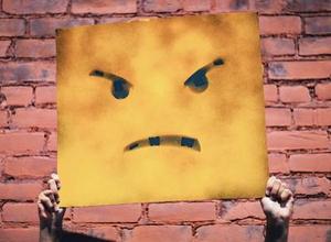 Vor einer Backsteinmauer wird ein Plakat mit einem wütenden Strichpunkte-Gesicht hochgehalten als Symbol für Hass