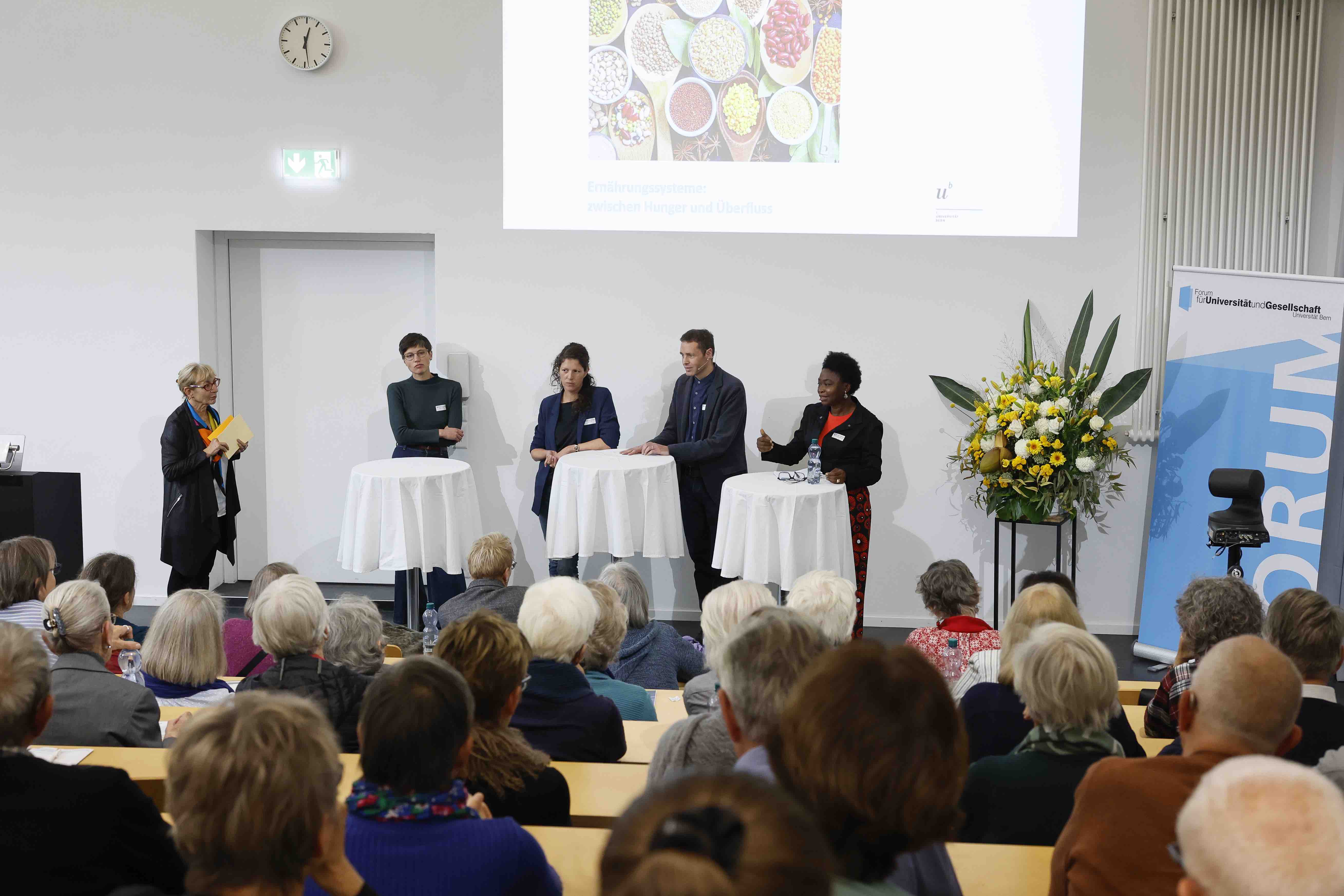 Ein voller Hörsaal, in dem das Publikum einer Podiumsdiskussion lauscht. Die Redner:innen stehen an Stehtischen mit weissen Tischtüchern.