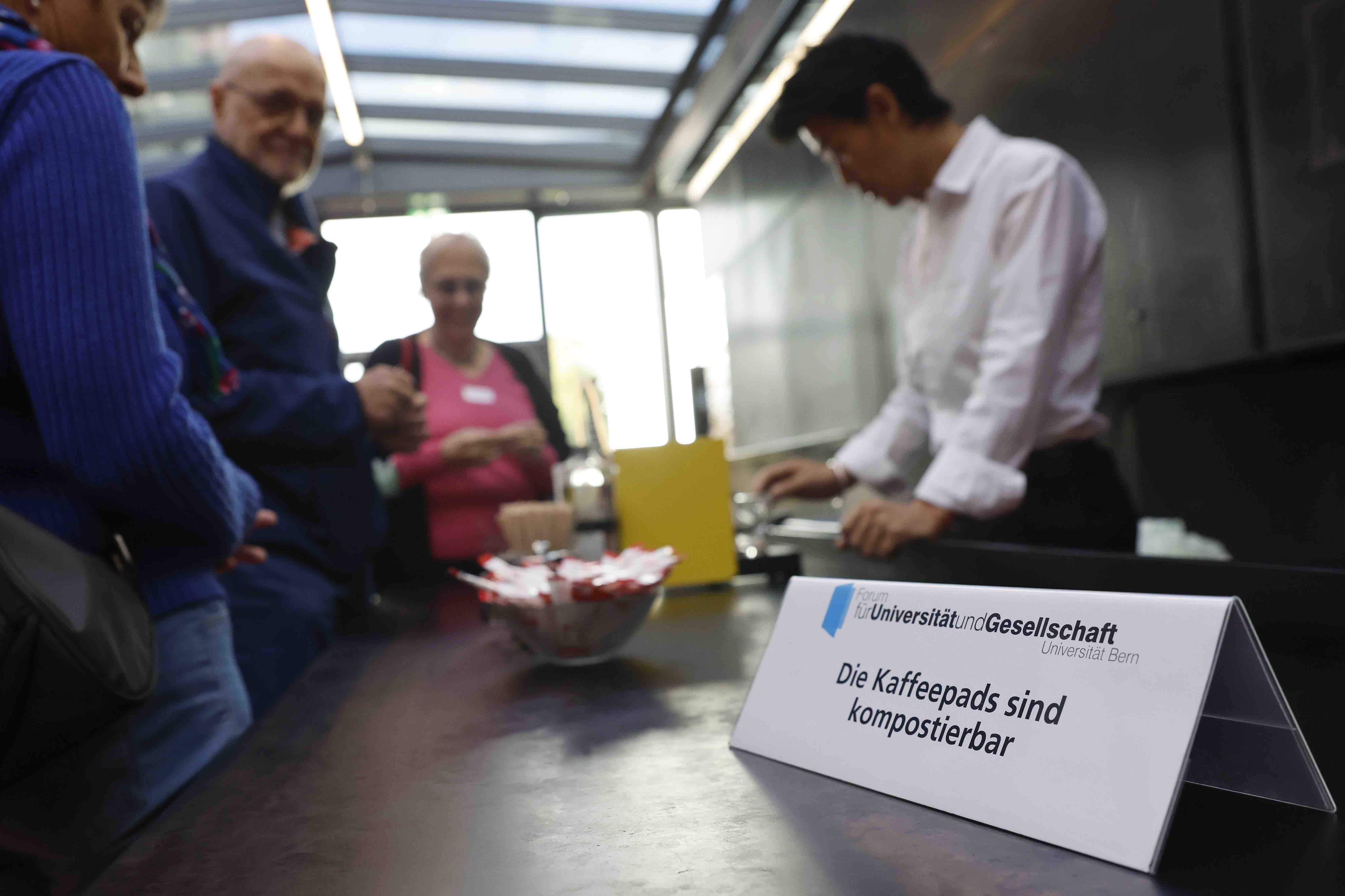 Aufnahme der Theke während eines Aperos. Im Vordergrund steht ein Schild mit dem Hinweis, dass die Kaffeepads kompostierbar sind.