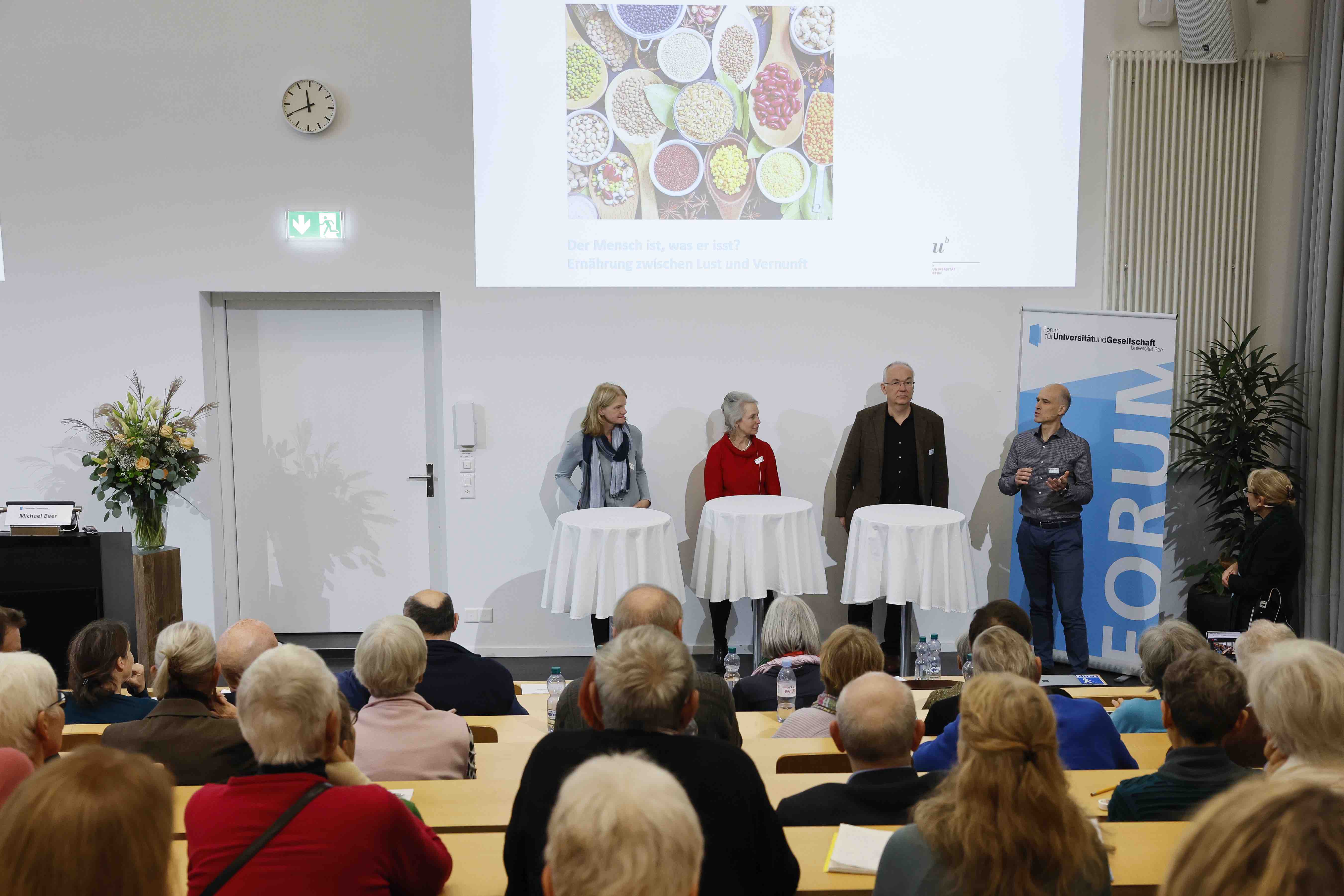 Drei Frauen und zwei Männer stehen an Stehtischen mit weissen Tischtüchern und diskutieren lachend mit dem Publikum, welches im Vordergrund zu sehen ist.