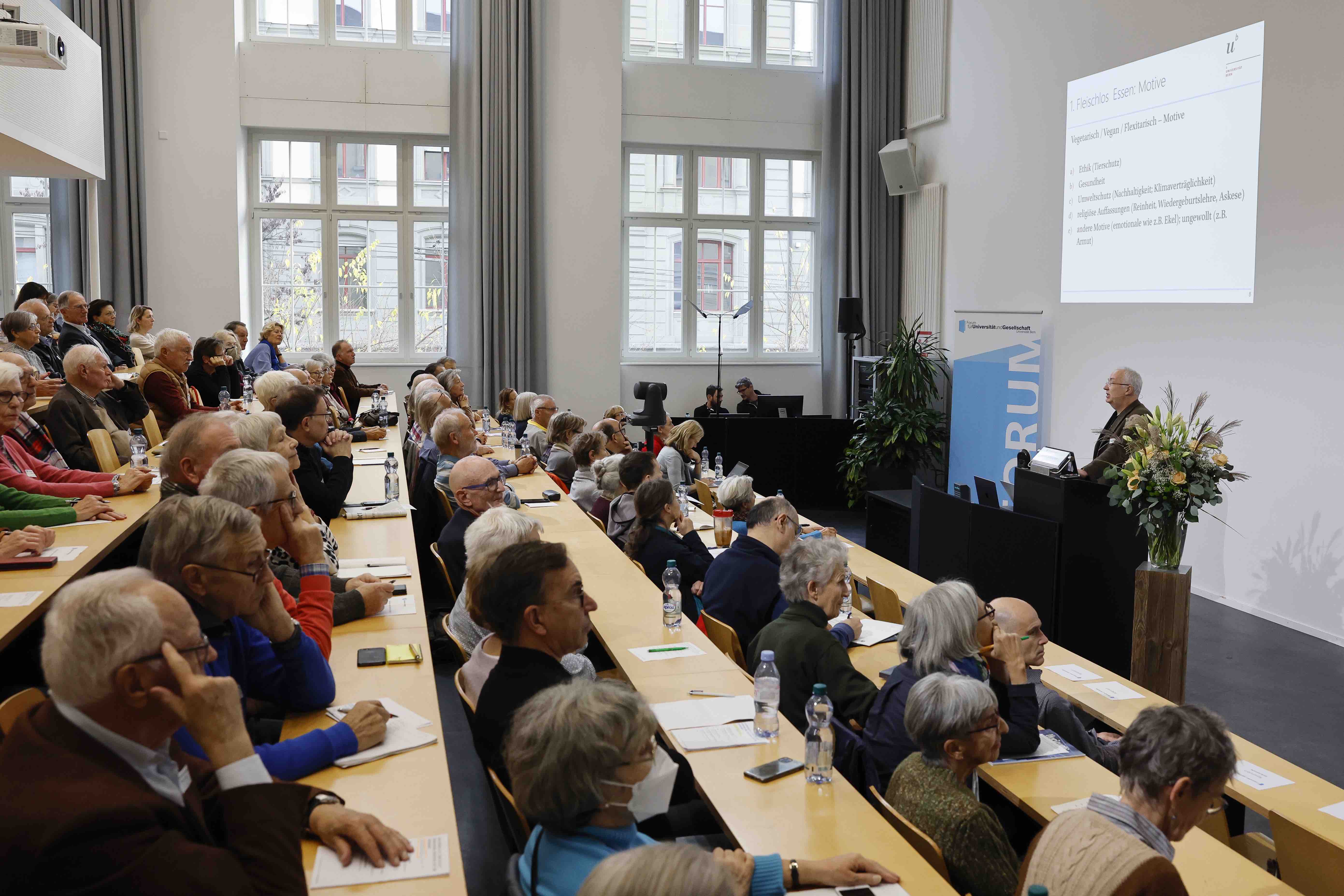 Blick von der Seite in einen gut besetzten Hörsaal. Das Publikum lauscht dem Referenten, der vorne am Rednerpult steht.