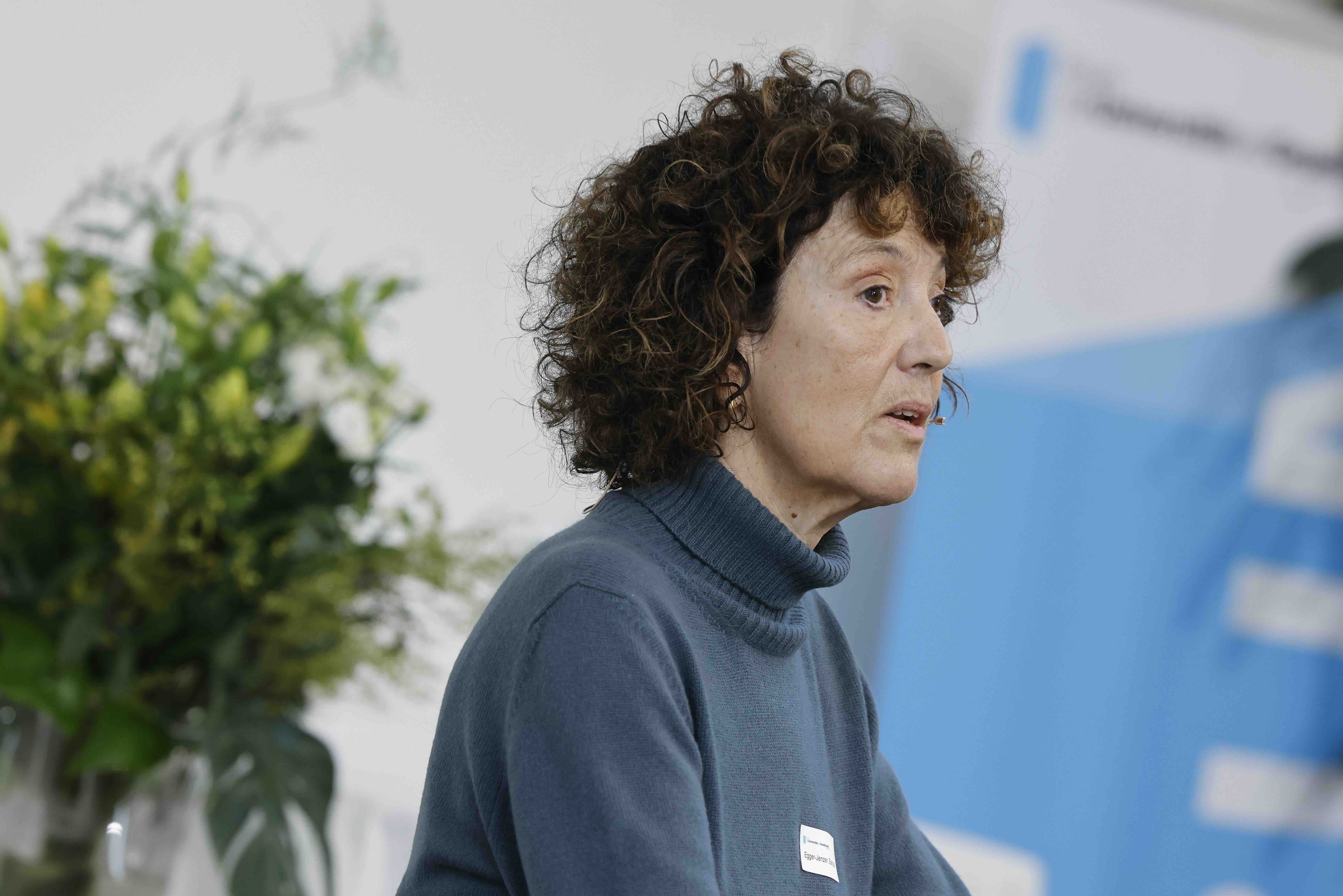 Eine Frau in einem petrolgrünen Rollkragenpullover ist zu sehen. Im Hintergrund snd ein Blumenstrauss und ein blau-weisses Banner zu sehen.