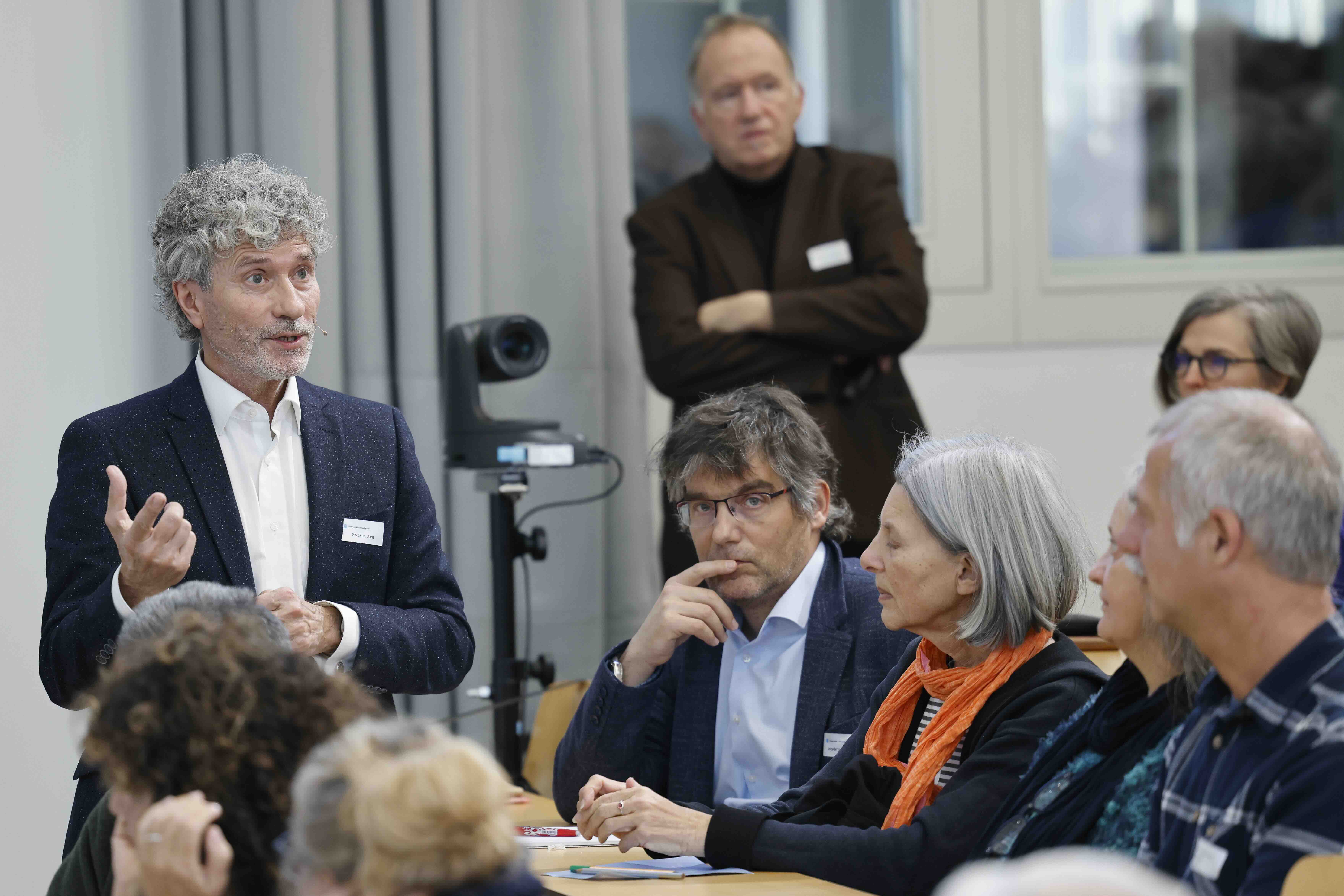 Ein Mann im Anzug diskutiert mit dem Publikum, welches im Vordergrund sichtbar ist.