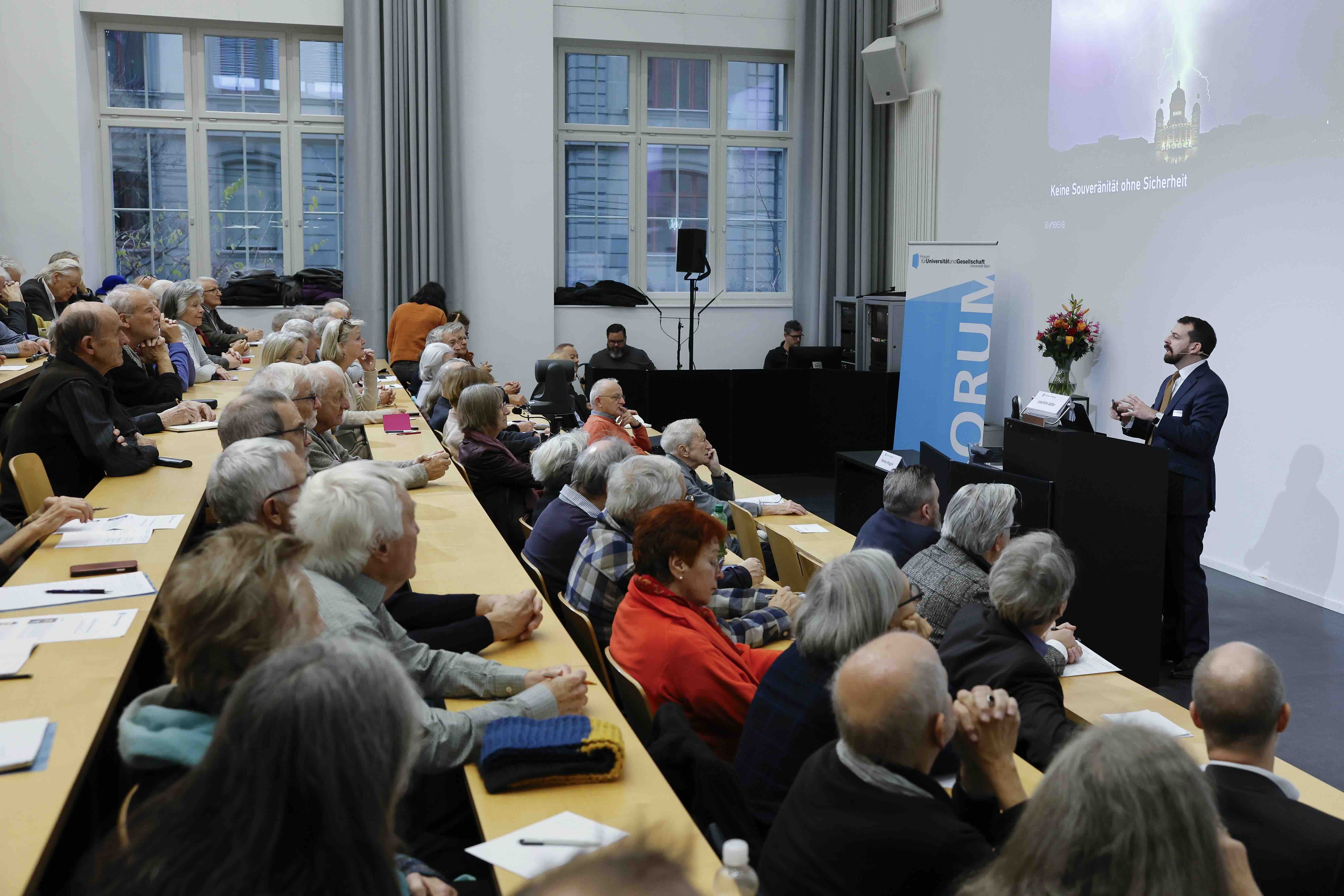 Eine Person steht vor vollen Reihen in einem Hörsaal an einem Rednerpult. Oberhalb von ihr ist die Projektion der Präsentation zu sehen, die ein Bild des Bundeshauses in einem Gewitter zeigt.