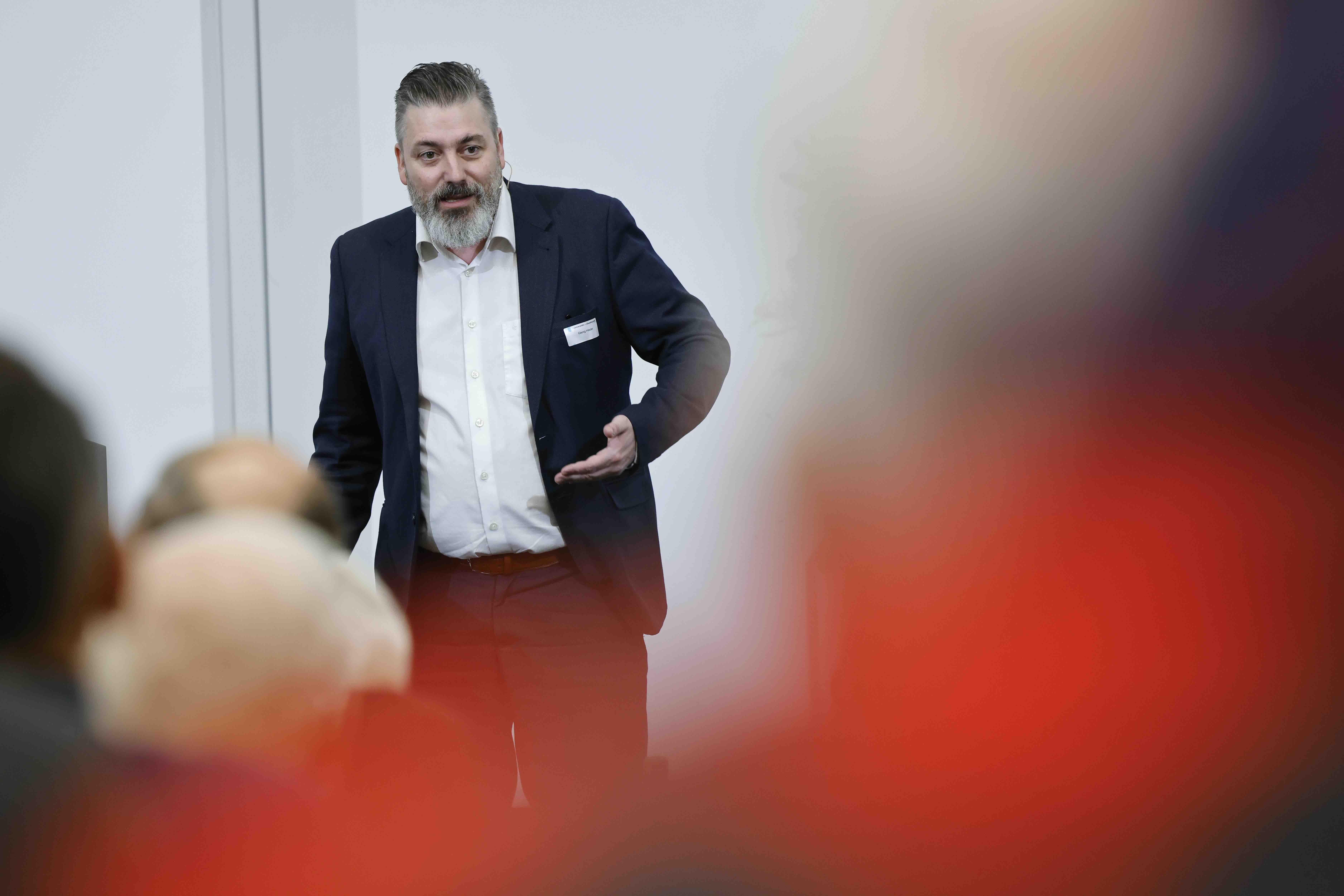Ein Mann mit Anzug und Bart spricht vor Publikum. Das Publikum ist verschwommen im Vordergrund erkennbar.