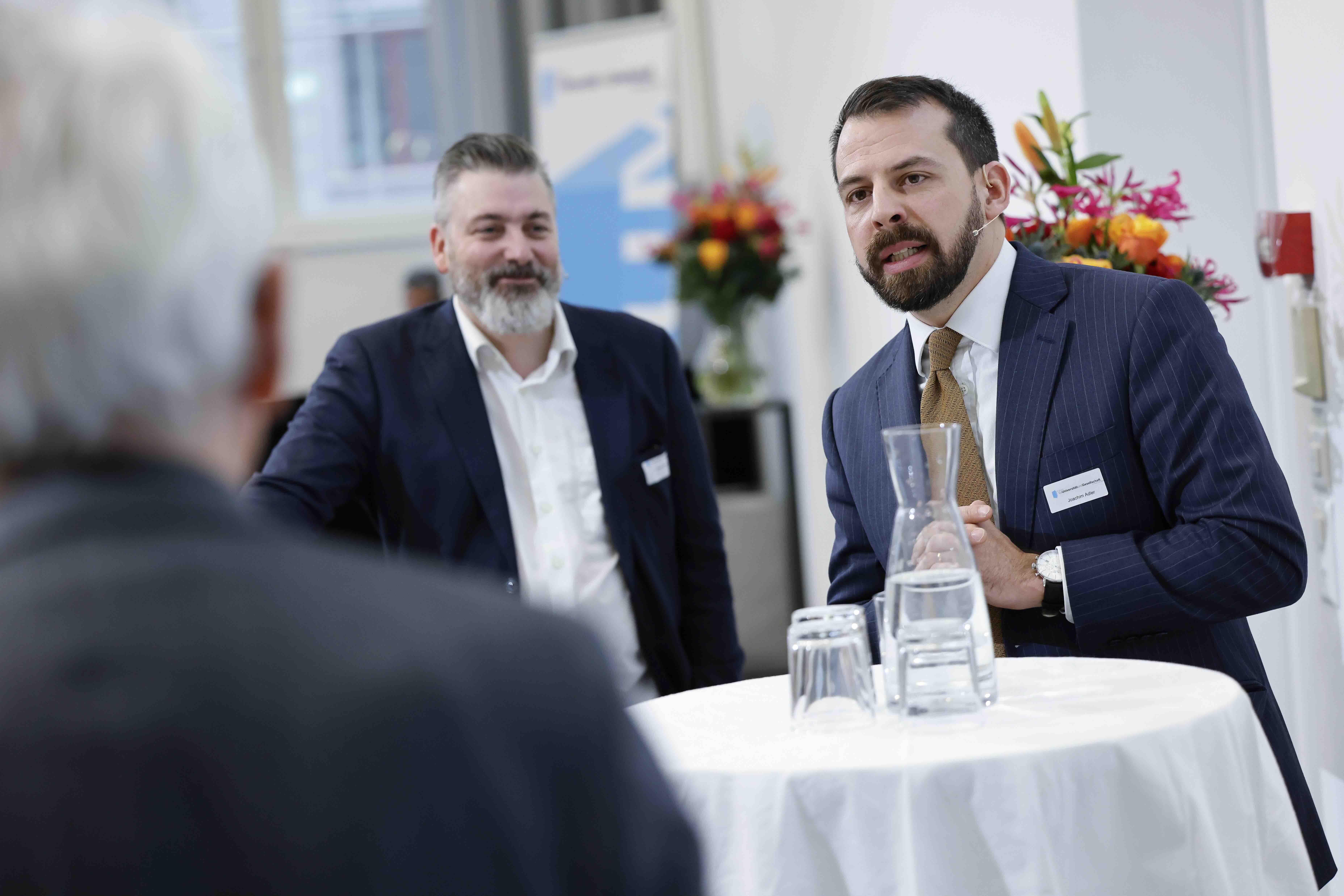 Zwei Männer im Anzug stehen an einem Stehtisch und diskutieren. Auf dem Tisch stehen eine Wasserkaraffe und Gläser und vorne links ist der Moderator von hinten zu erkennen.