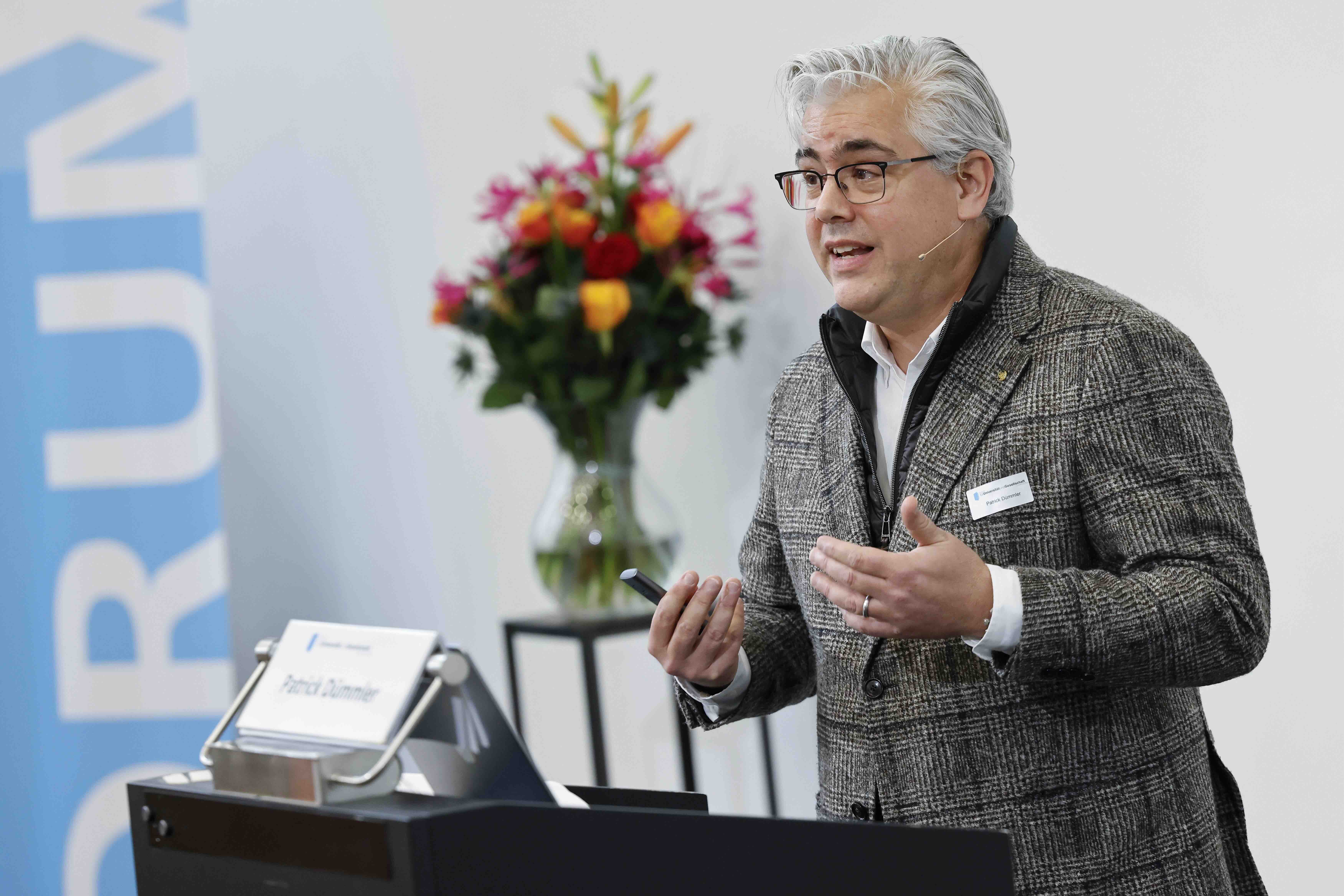 Ein Mann mit Brille und grauem Jackett steht hinter einem Rednerpult und gestikuliert mit beiden Händen. In seiner rechten Hand hält er einen Presenter. Im Hintergrund sind verschwommen ein blau-weisses Banner und ein bunter Blumenstrauss sichtbar.