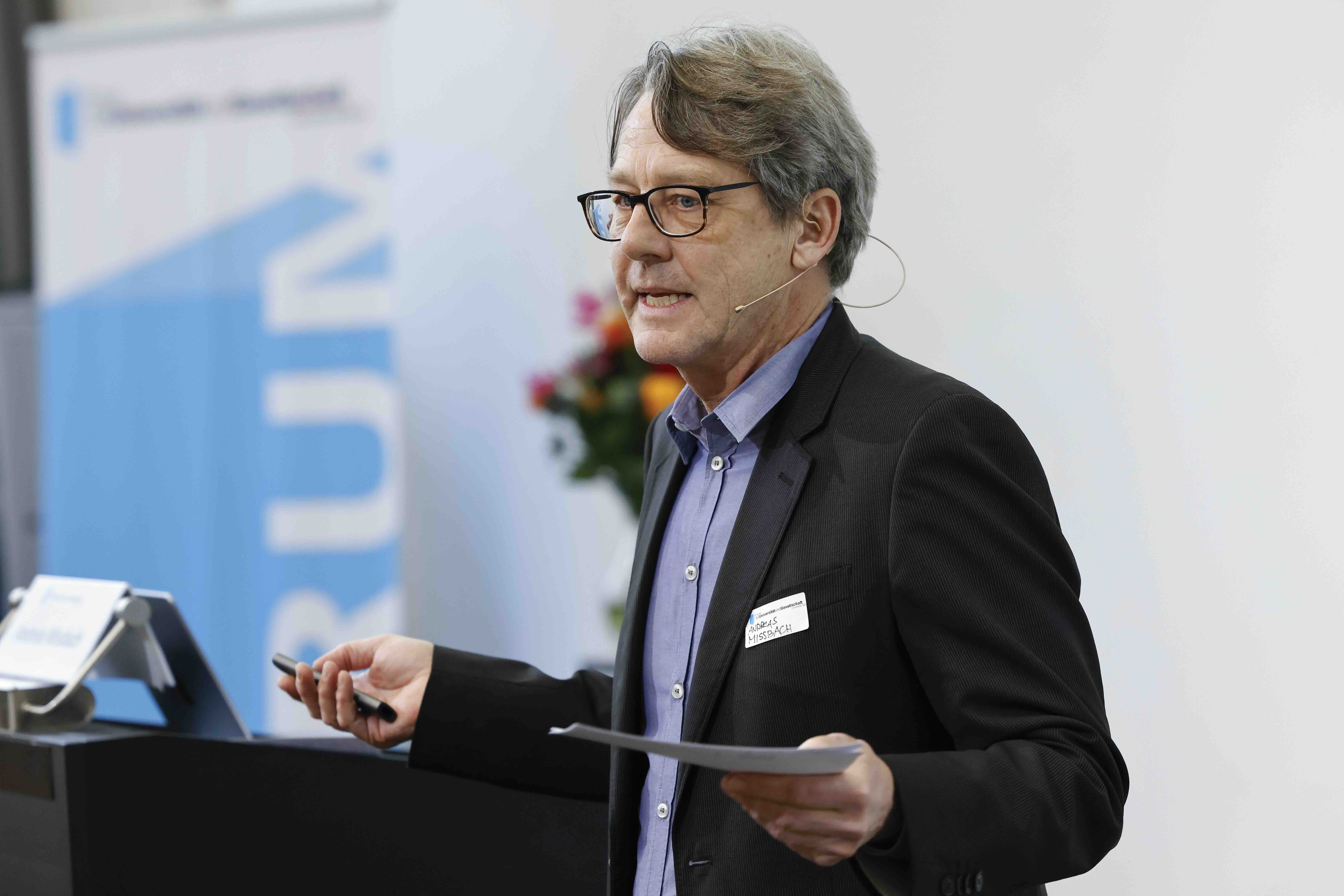 Ein Mann mit Brille und dunklem Anzug steht neben einem Rednerpult. in seiner rechten Hand hält er einen Presenter, in der linken sein Manuskript. Im Hintergrund sind ein blau-weisses Banner und ein farbiger Blumenstrauss erkennbar.
