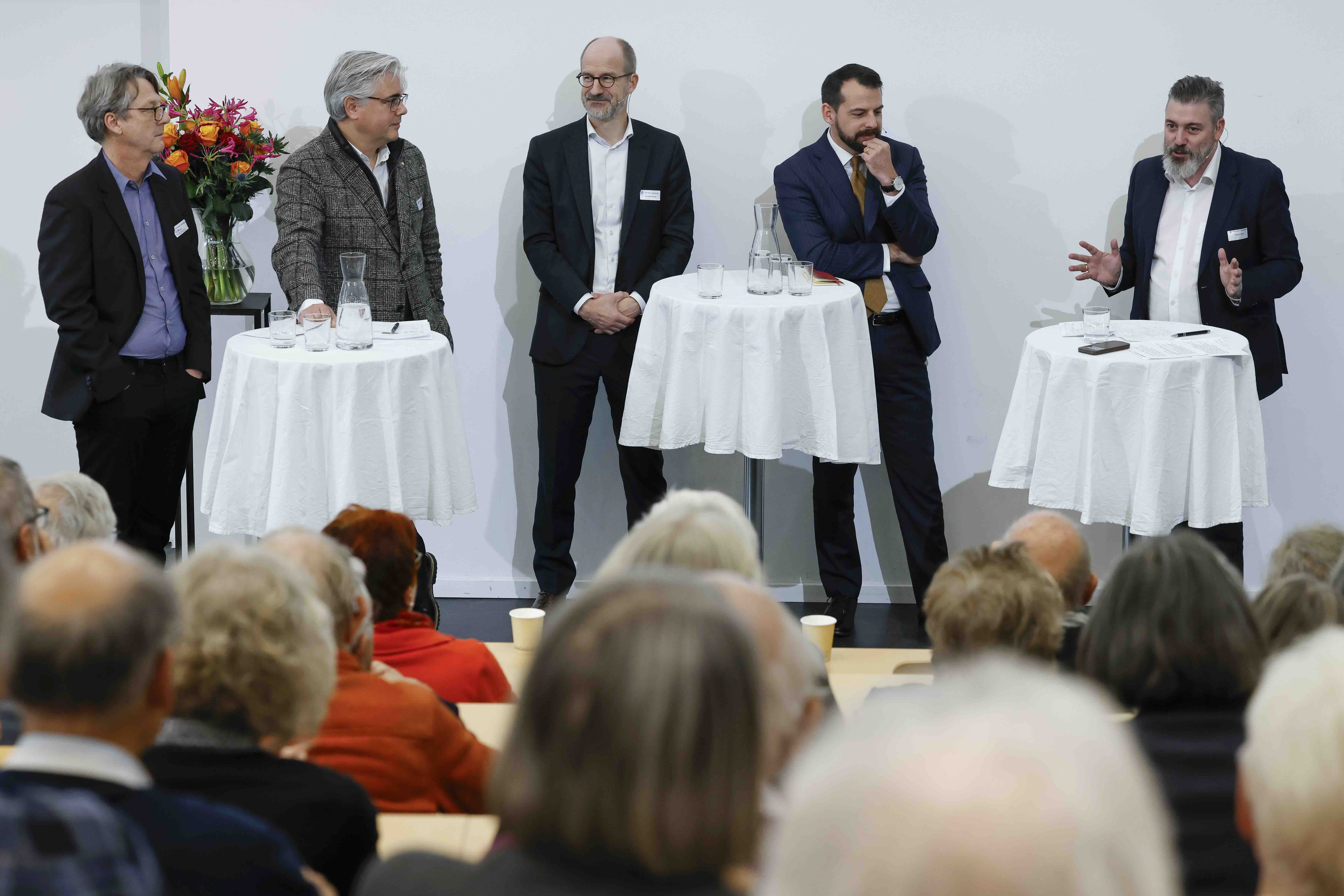 Fünf Männer im Anzug stehen hinter drei Stehtischen mit weissen Tischtüchern. Auf den Tischen stehen Gläser und eine Wasserkaraffe. Der Mann rechts im Bild ist am Sprechen und gestikuliert mit beiden Händen, die anderen hören zu.
