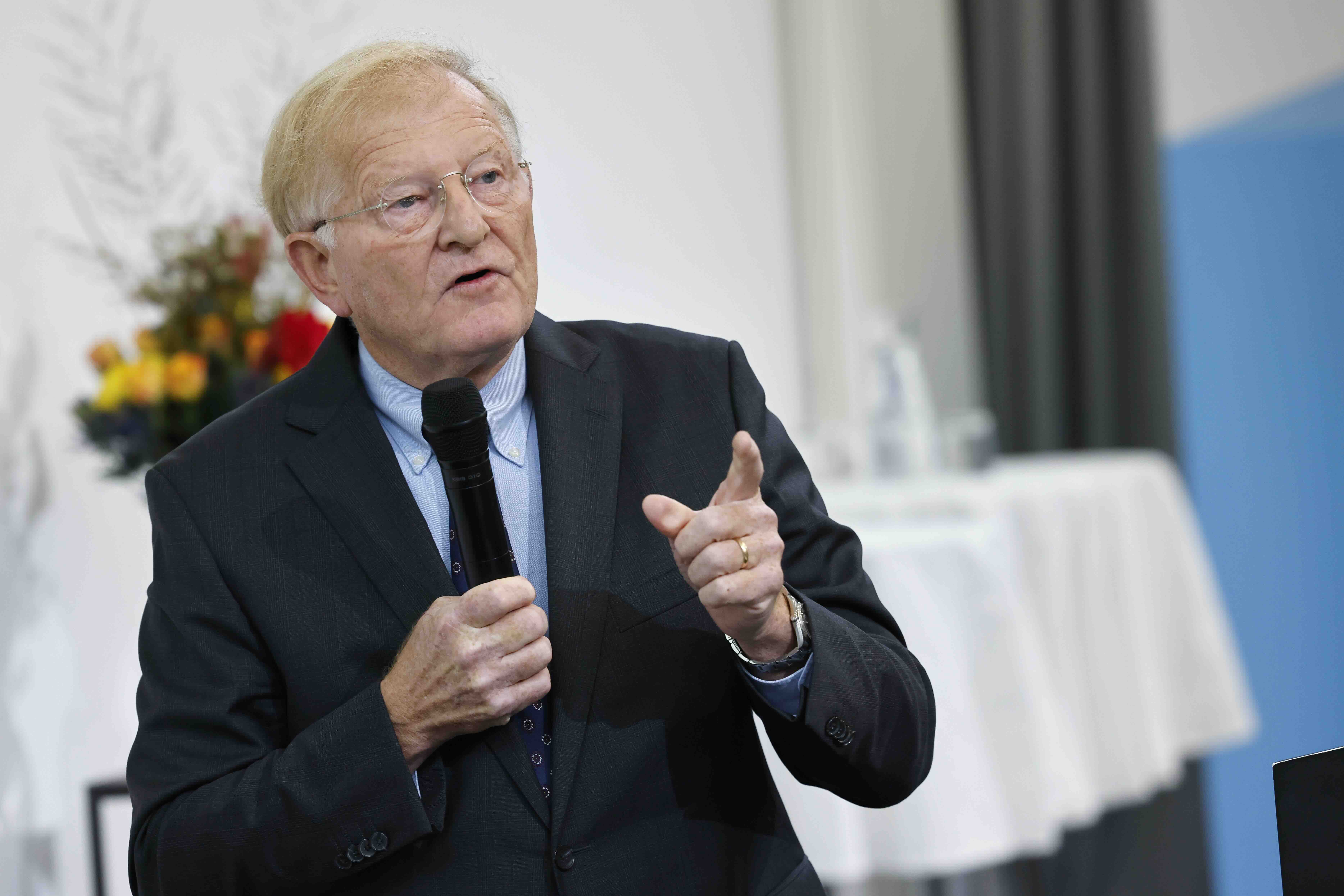 Thomas Cottier trägt einen Anzug mit dunkler Krawatte. Er hält in der rechten Hand ein Mikrofon und gestikuliert mit der linken. Hinter ihm sind ein rot-gelber Blumenstrauss und Stehtische mit weissen Tischtüchern erkennbar.