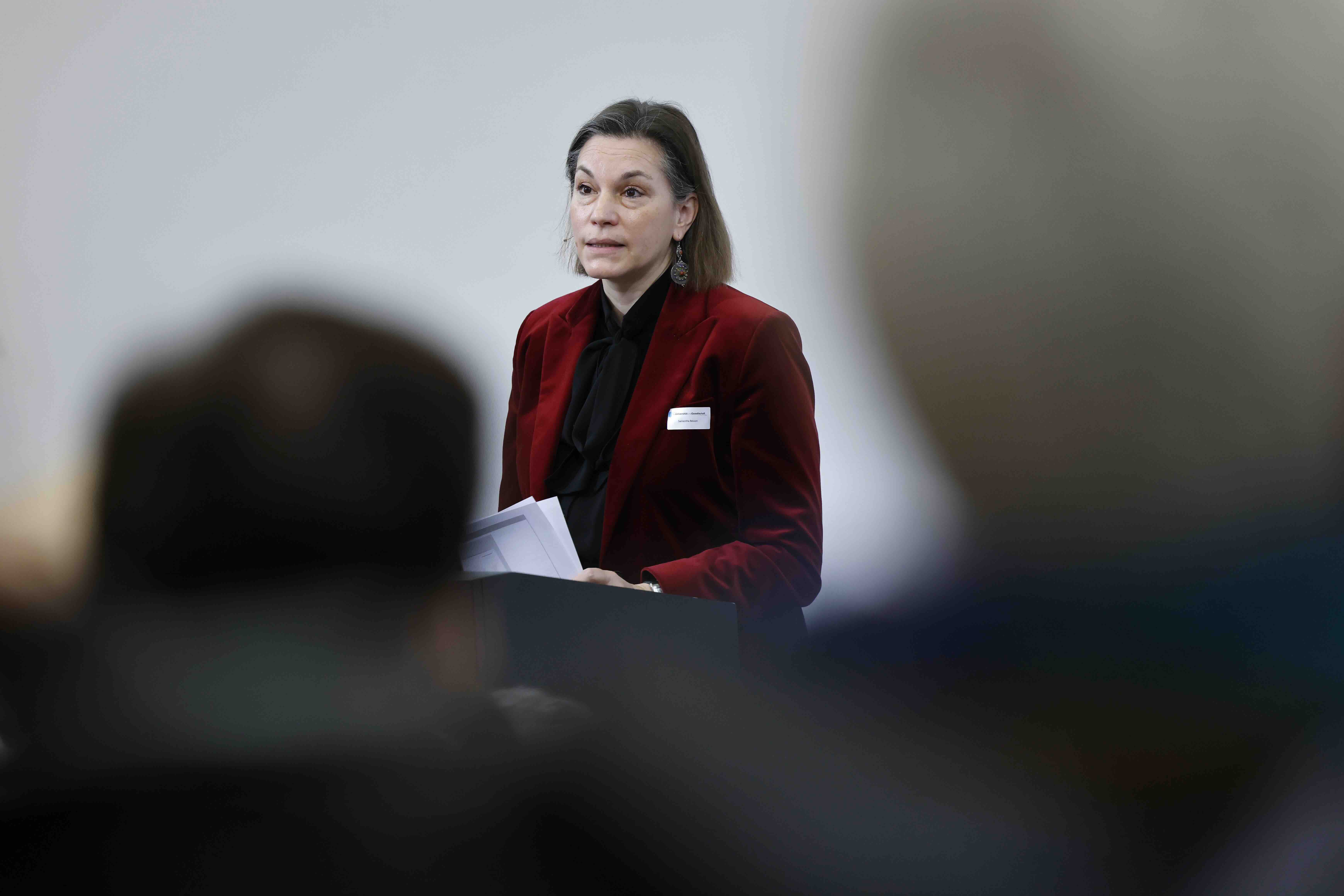 Samantha Besson trägt ein rotes Jackett. Sie steht hinter einem Rednerpult und hält ein Manuskript in den Händen. Im Vordergrund ist verschwommen ein Teil des Publikums erkennbar.