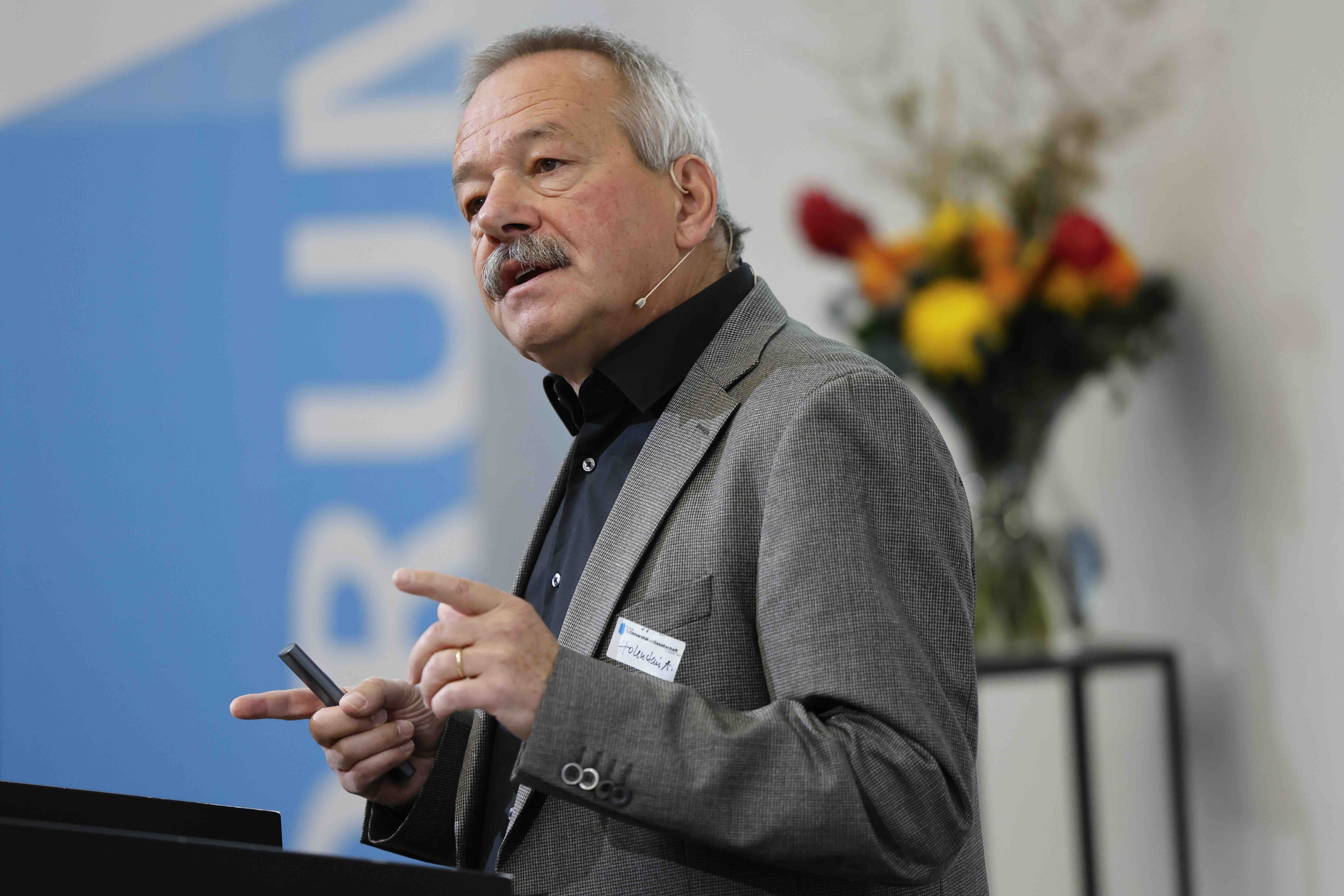 André Holenstein trägt ein graues Jackett und ein dunkles Hemd. Er steht hinter einem Rednerpult ud gestikuliert mit beiden Händen, in der rechten hält er einen Presenter. Im Hintergrund sind ein rot-gelber Blumenstrauss und ein blau-weisses Banner erkennbar.