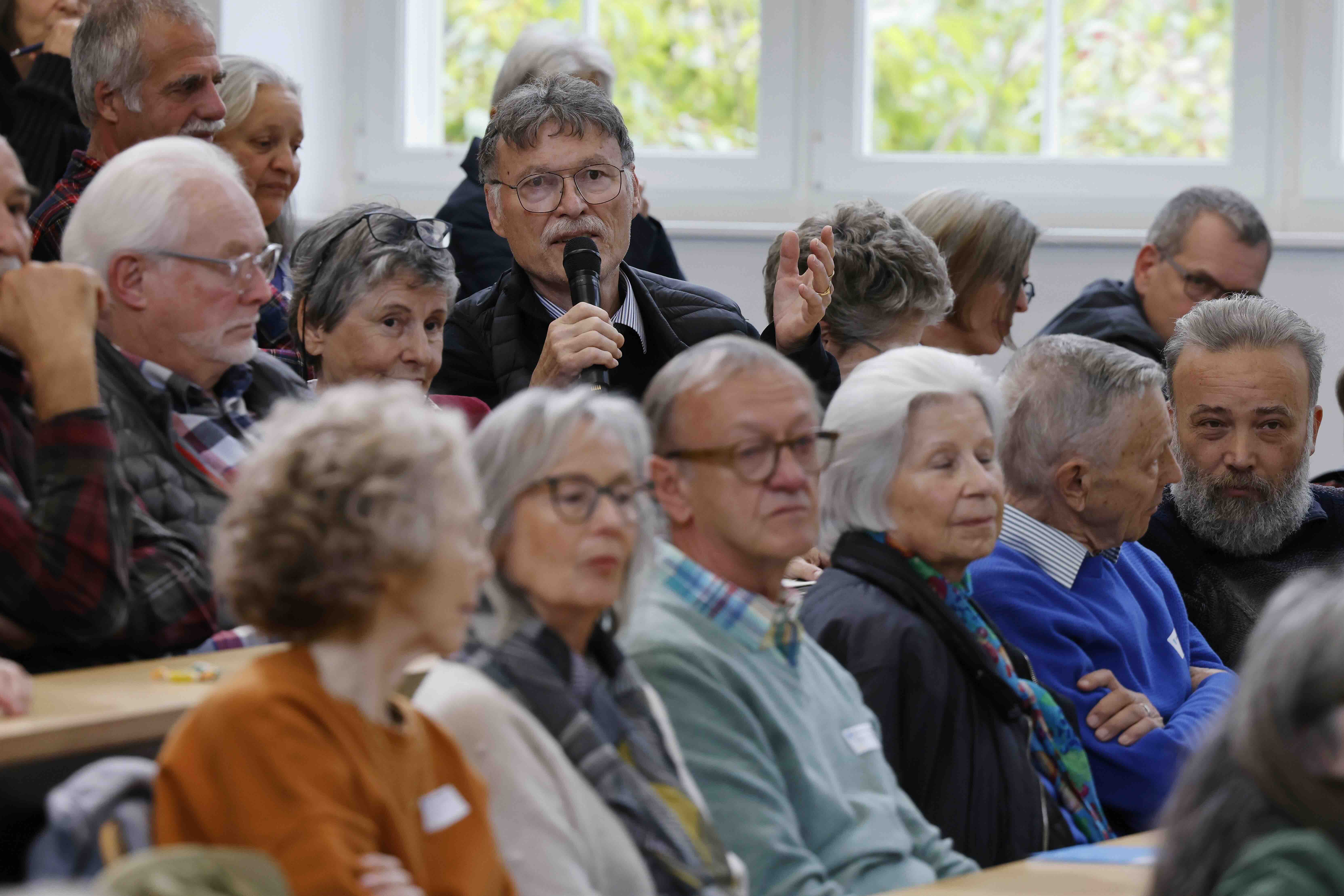 Das Bild zeigt einen Ausschnitt des Publikums, in der oberen Bildhälfte ist ein Mann sichtbar, der in ein Mikrofon spricht.
