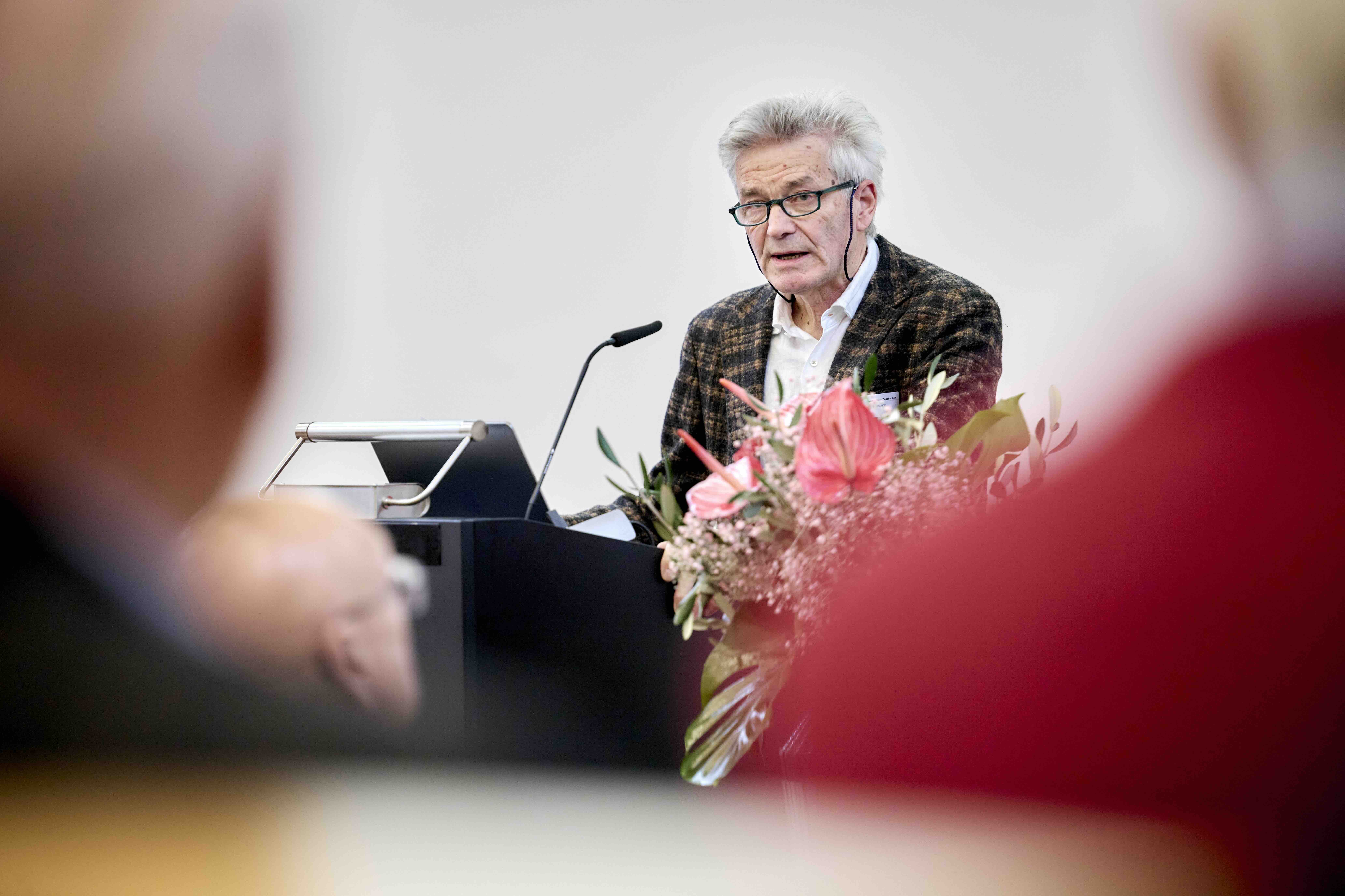 Ein älterer Mann mit Brille spricht an einem Rednerpult bei einem öffentlichen Vortrag; im Vordergrund sind unscharf sitzende Zuhörende und ein Blumenarrangement zu sehen.