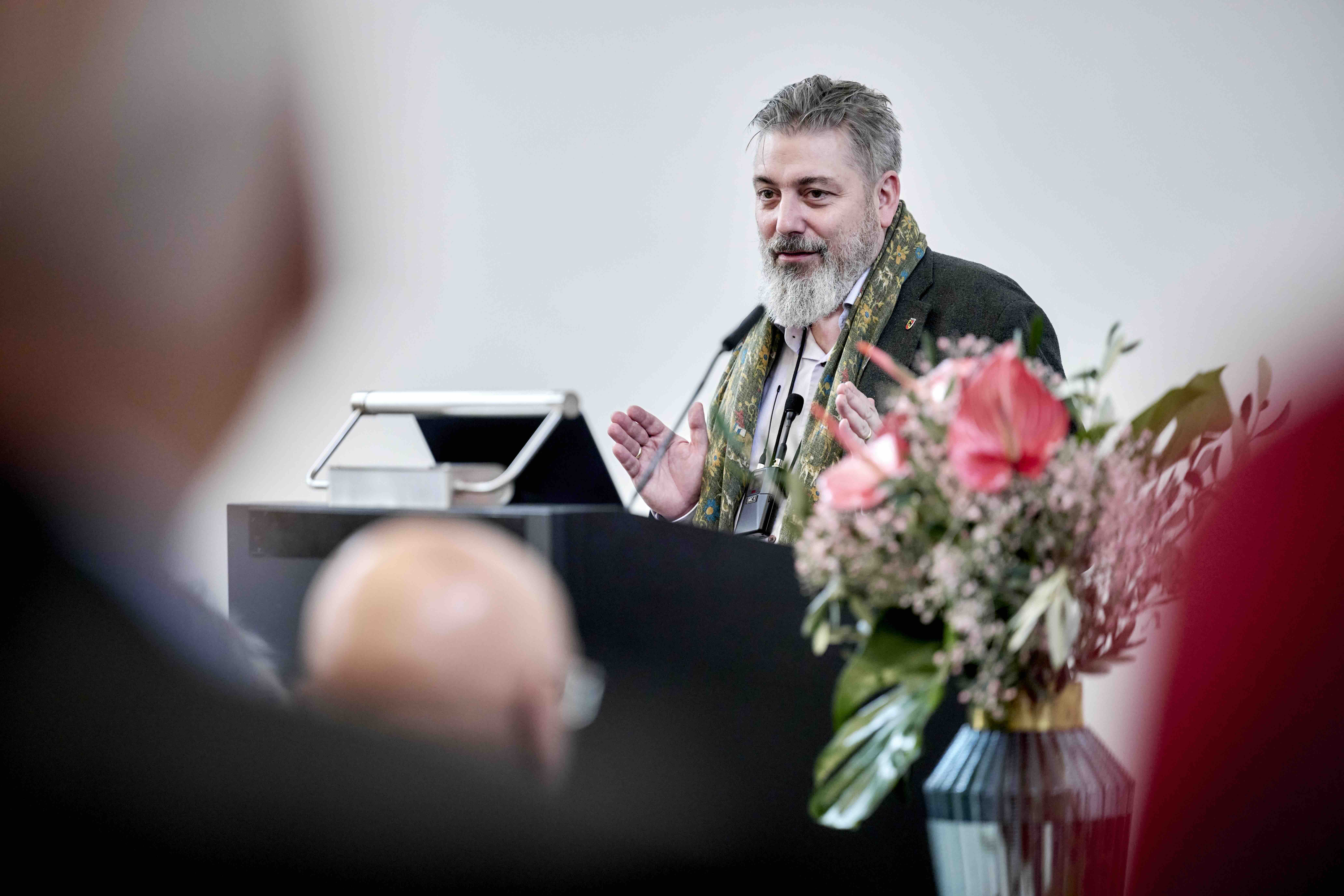 Ein Mann mit grauem Bart spricht an einem Rednerpult bei einem öffentlichen Vortrag und gestikuliert mit den Händen; im Vordergrund sind unscharf Zuhörende und ein Blumenarrangement zu sehen.