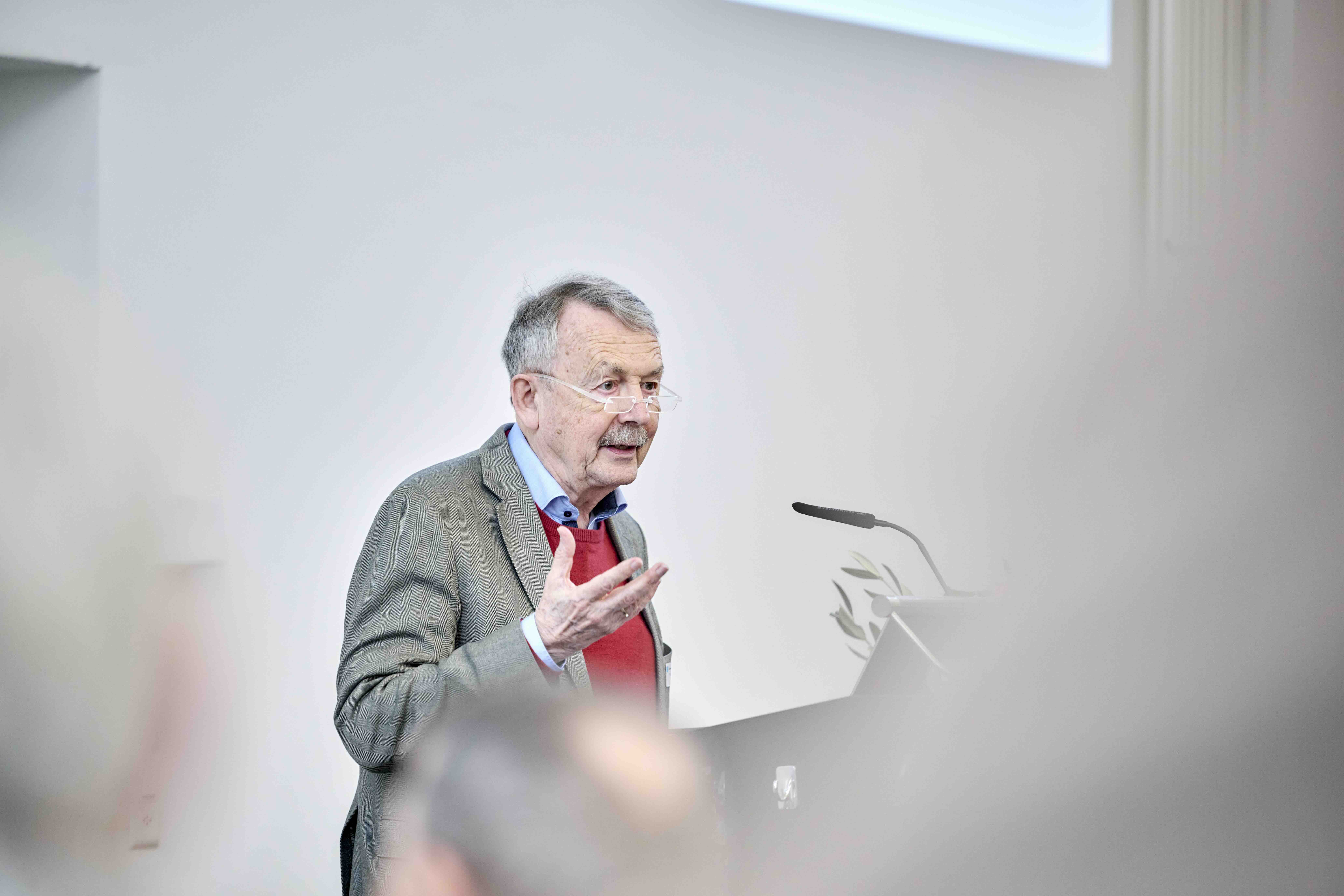 Ein älterer Mann mit Brille hält einen Vortrag an einem Rednerpult und gestikuliert mit einer Hand; im Vordergrund sind unscharf sitzende Zuhörende zu erkennen.