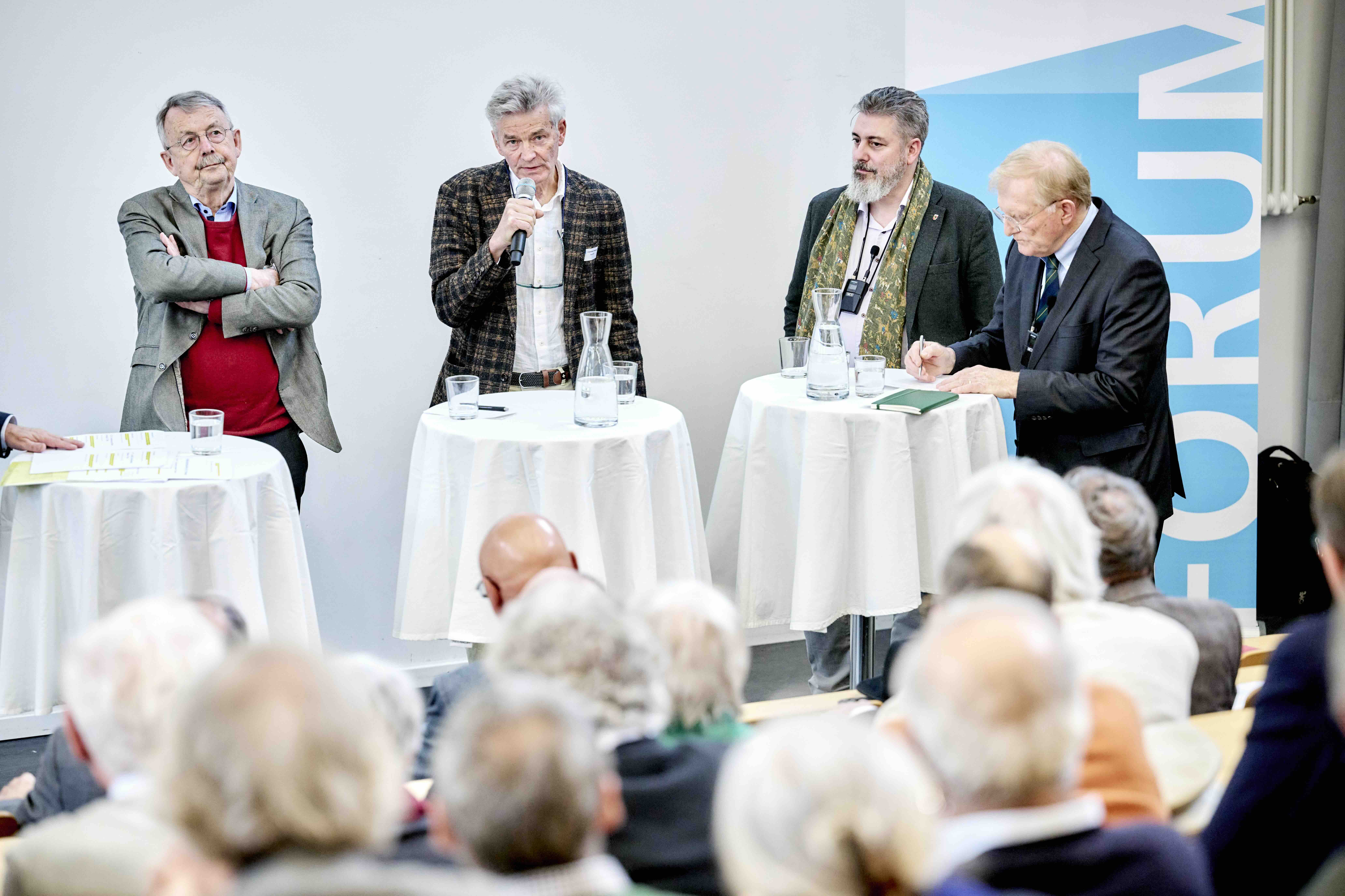 Vier Männer stehen an Stehtischen auf einem Podium bei einer öffentlichen Diskussion; einer spricht ins Mikrofon, andere hören zu oder machen Notizen, im Vordergrund sitzt das Publikum.