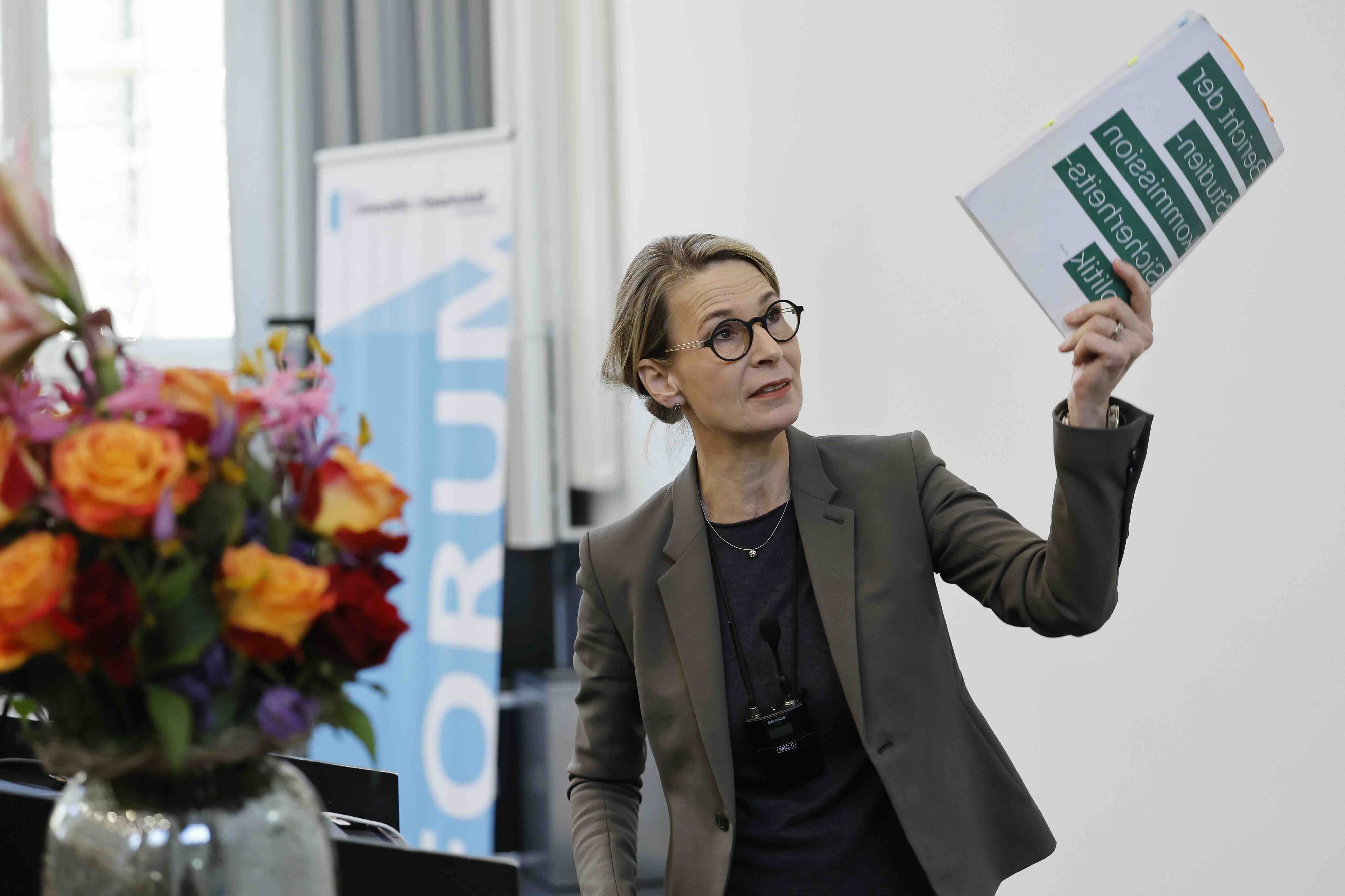 Eine Frau mit Brille und zusammengebundenen Haaren steht an einem Rednerpult und hält ein Dokument mit grünen Textfeldern hoch. Sie trägt einen dunklen Blazer und spricht vor einem hellen Hintergrund. Links im Bild steht ein bunter Blumenstrauß in einer Glasvase, im Hintergrund ist ein Banner des Forums für Universität und Gesellschaft sichtbar.