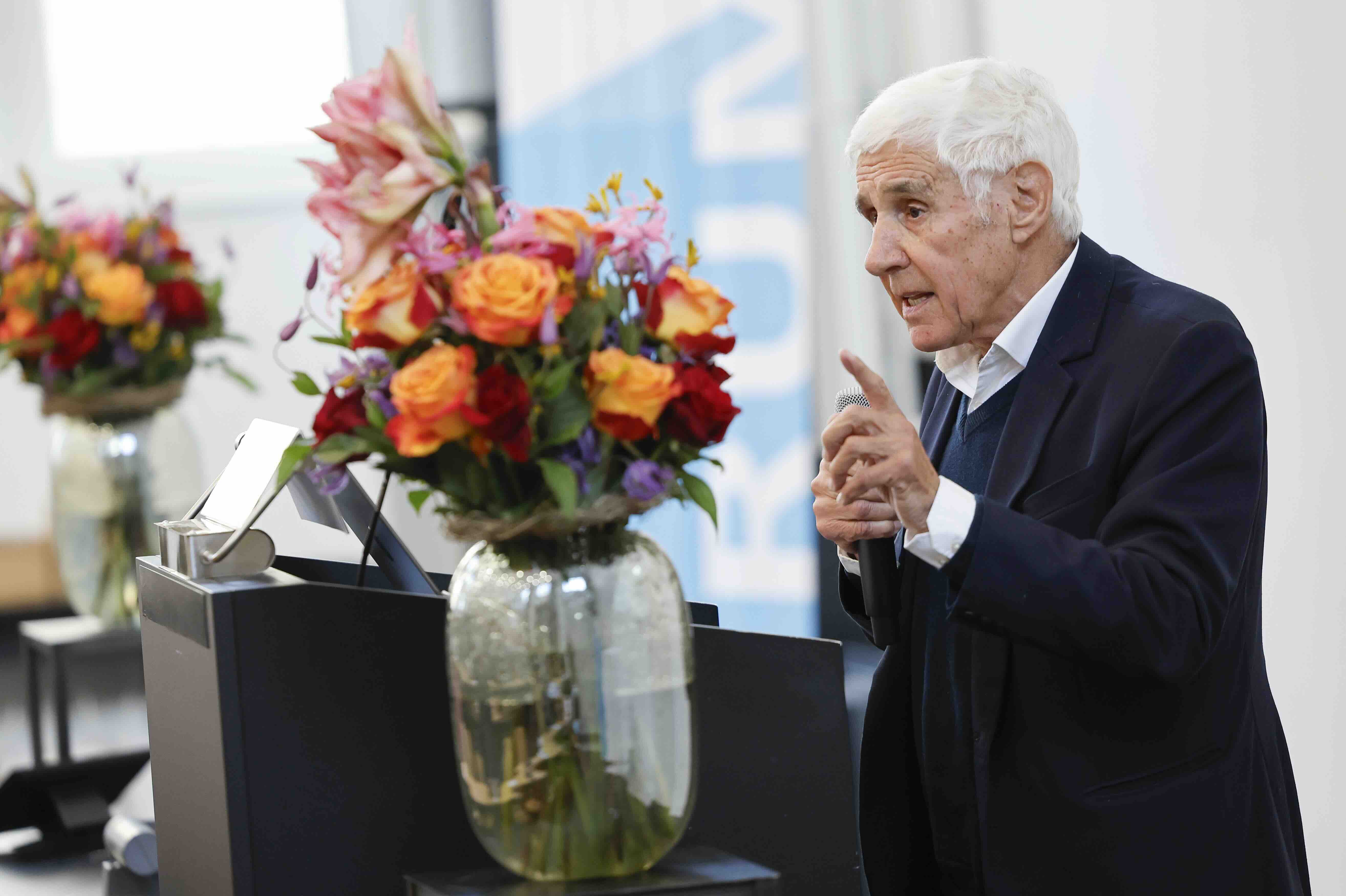 Ein älterer Mann mit weißem Haar spricht bei einem Vortrag und hält ein Mikrofon in der Hand. Er trägt ein dunkles Jackett und gestikuliert mit erhobenem Finger. Vor ihm steht ein großer bunter Blumenstrauß in einer Glasvase, der den Vordergrund prägt. Der Hintergrund ist hell und zeigt ein unscharfes Banner.