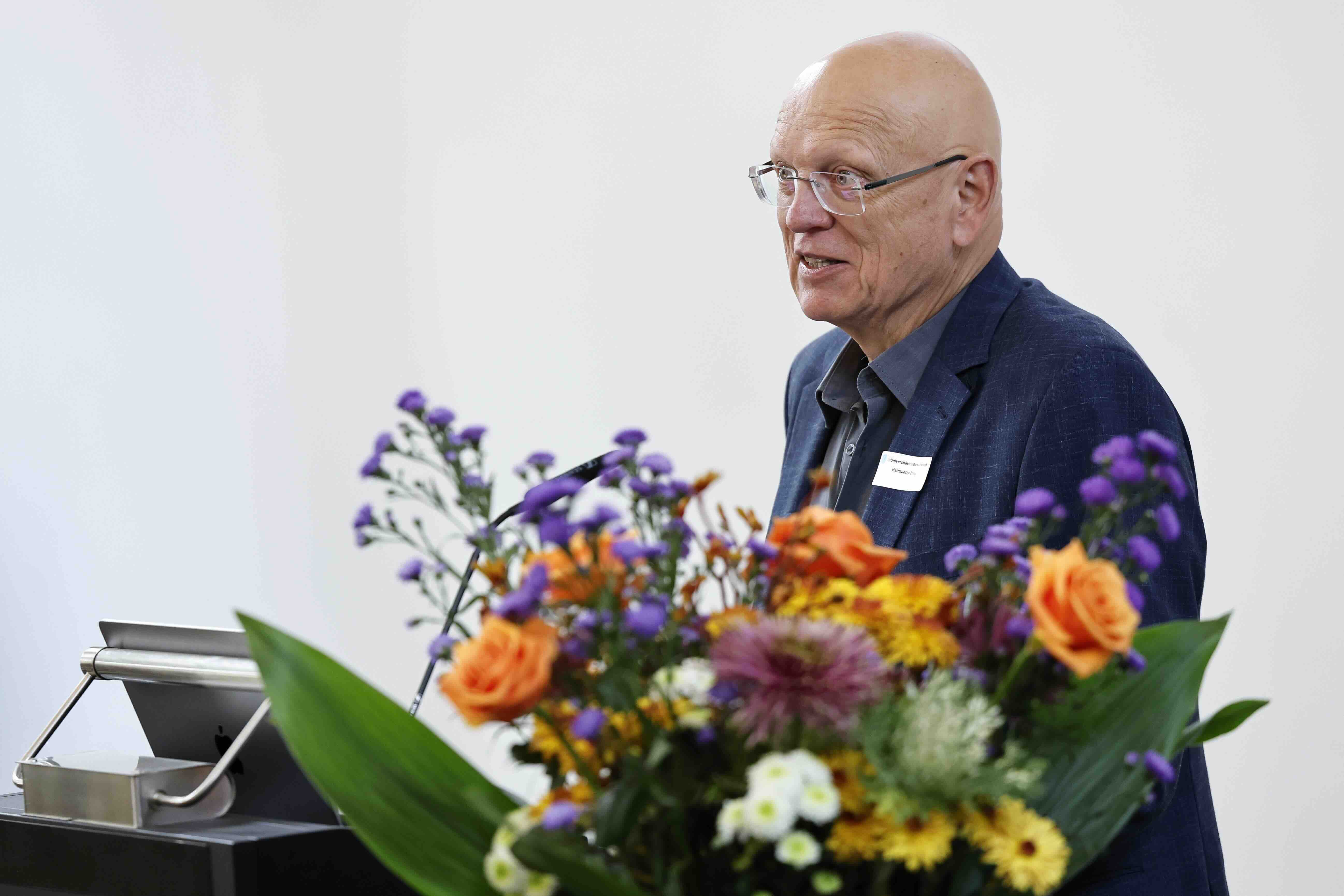 Ein älterer Mann mit Glatze und Brille steht an einem Rednerpult und spricht. Er trägt ein blaues Sakko und wirkt konzentriert. Im Vordergrund steht ein großer, bunter Blumenstrauß mit orangen Rosen und weiteren farbigen Blüten