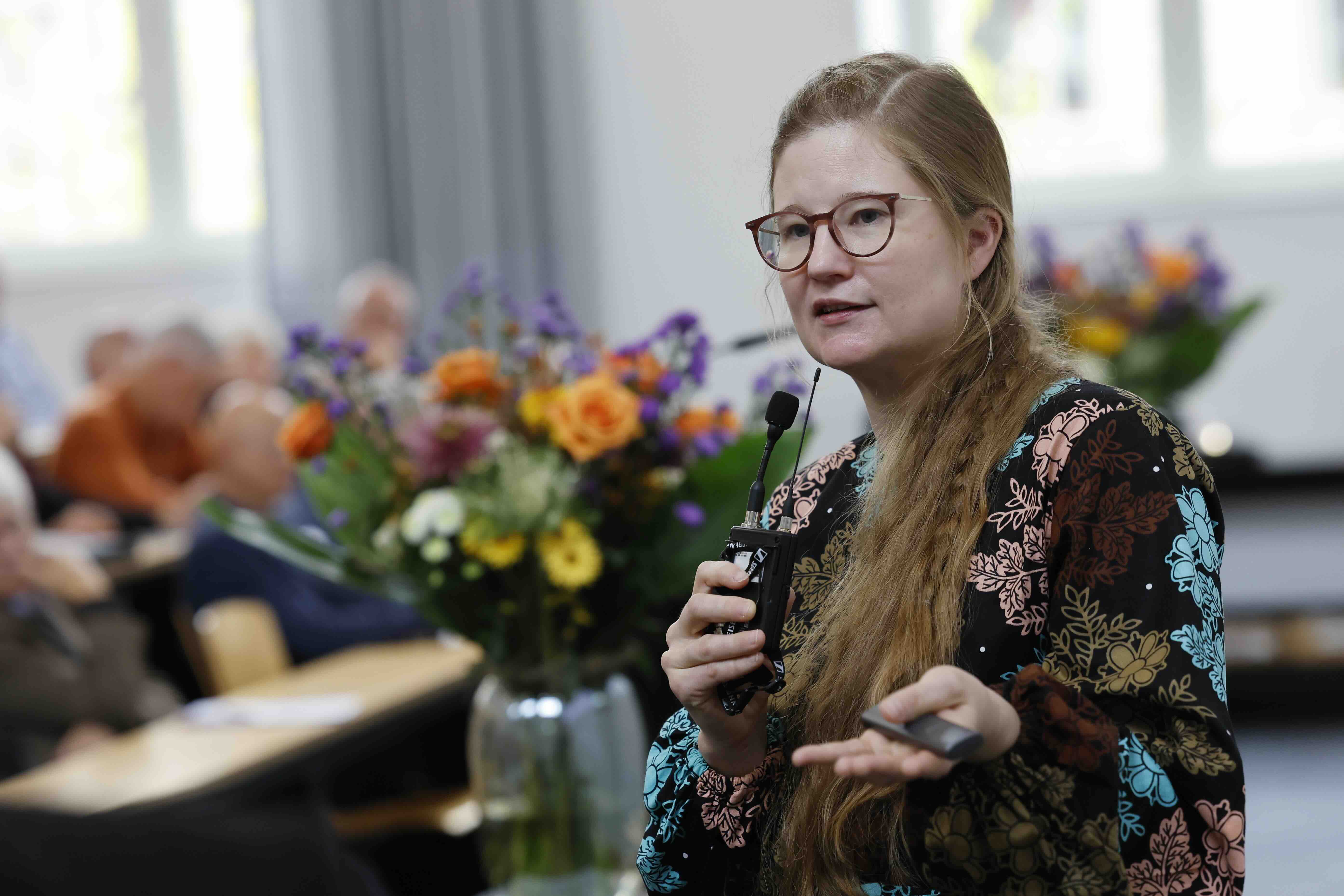 Auf dem Bild ist eine Frau in einem Vortragsraum zu sehen. Sie spricht vor Publikum und hält ein kabelloses Mikrofon sowie einen Präsentationsklicker in den Händen. Die Frau trägt eine Brille und ein bunt gemustertes Kleid, ihre langen Haare sind über die Schulter gelegt. Im Hintergrund sitzen mehrere Zuhörende und auf einem Tisch stehen große Blumensträuße in Vasen.