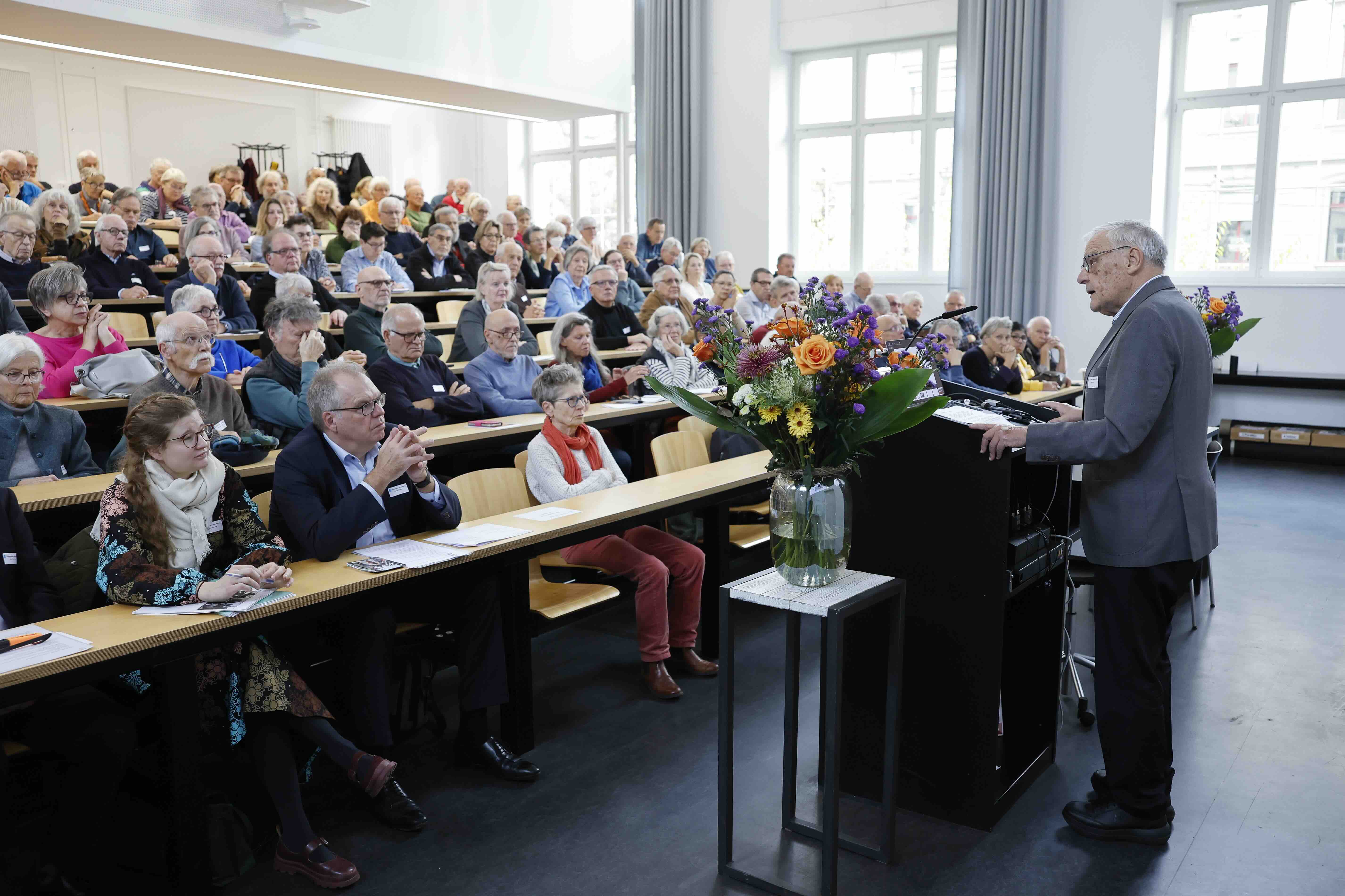 In einem gut besuchten Hörsaal spricht ein älterer Mann am Rednerpult zu einem grossen Publikum. Die Zuhörenden sitzen in mehreren Reihen und hören aufmerksam zu. Im Raum steht Tageslicht durch grosse Fenster. Auf dem Pult und im Raum stehen Blumensträusse mit bunten Schnittblumen.