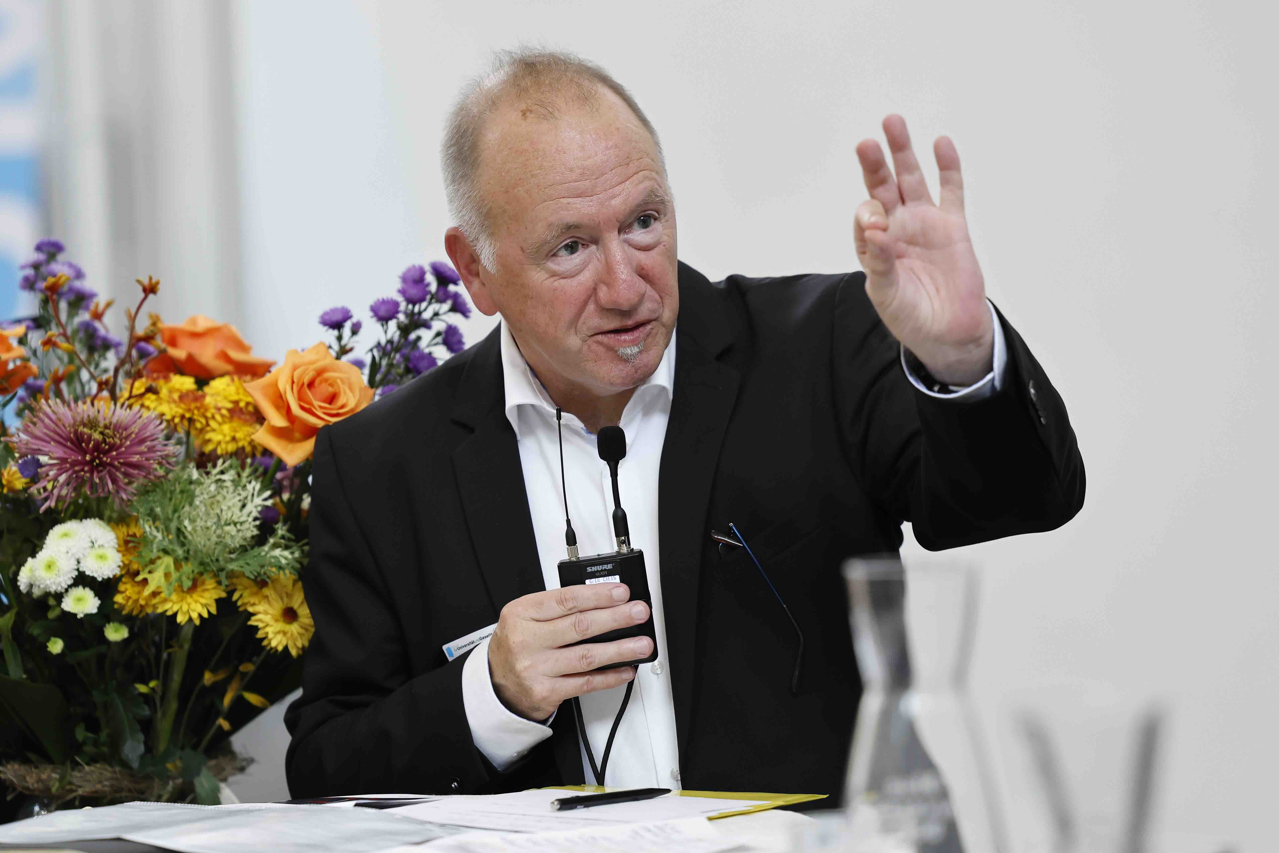 Ein Mann in dunklem Anzug spricht in ein Mikrofon und gestikuliert mit erhobener Hand. Er sitzt an einem Tisch mit Unterlagen, im Hintergrund stehen bunte Blumenarrangements.