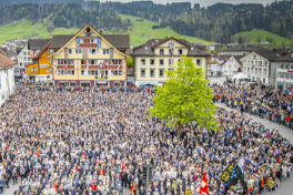 Grosse Menschenmenge bei einer traditionellen Landsgemeinde auf dem Dorfplatz von Appenzell, umgeben von historischen Gebäuden und grüner Hügellandschaft; zentral steht ein großer Baum, zahlreiche Teilnehmende versammeln sich zur öffentlichen Abstimmung im Freien.