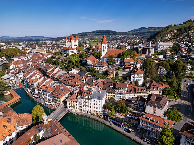 Luftaufnahme der Thuner Altstadt mit Aare, Schloss Thun, Stadtkirche und dicht bebautem Stadtzentrum vor der hügeligen Landschaft des Berner Oberlands.