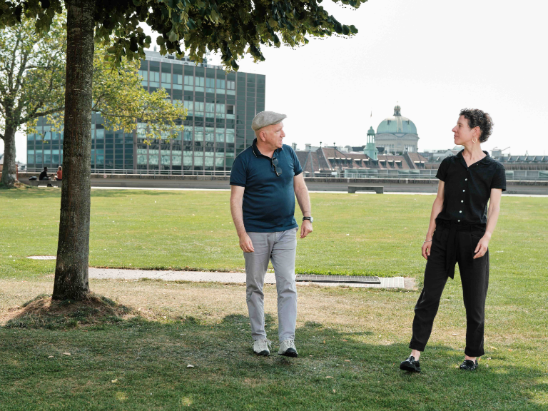 Zwei Personen stehen auf der Grossen Schanze in Bern im Gespräch. Im Hintergrund sind das Bundeshaus und ein modernes Bürogebäude sichtbar, im Vordergrund ein Baum und eine Rasenfläche.