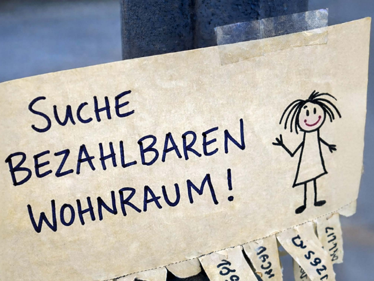 Handgeschriebener Zettel mit der Aufschrift "Suche bezahlbaren Wohnraum!" neben einer einfachen Kinderzeichnung einer Person; im Hintergrund sind unscharfe Wohnungsanzeigen zu sehen. Das Bild symbolisiert die dringende Suche nach günstigem, bezahlbaren Wohnraum und thematisiert die aktuelle Wohnungsnot.