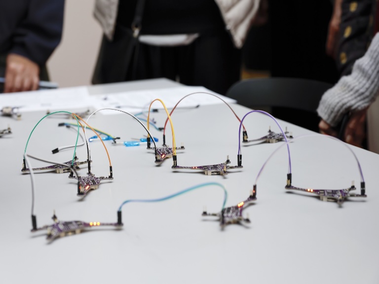 Mehrere kleine elektronische Platinen in Sternform mit LEDs sind über bunte Kabel auf einem Tisch verbunden. Sie simulieren ein künstliches neuronales Netzwerk zur Veranschaulichung von Künstlicher Intelligenz. Personen stehen im Hintergrund.