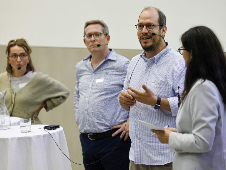 Vier Personen mit Mikrofonen im Gespräch bei einer Podiumsdiskussion. Eine Frau moderiert, während zwei Männer und eine Frau zuhören und diskutieren.