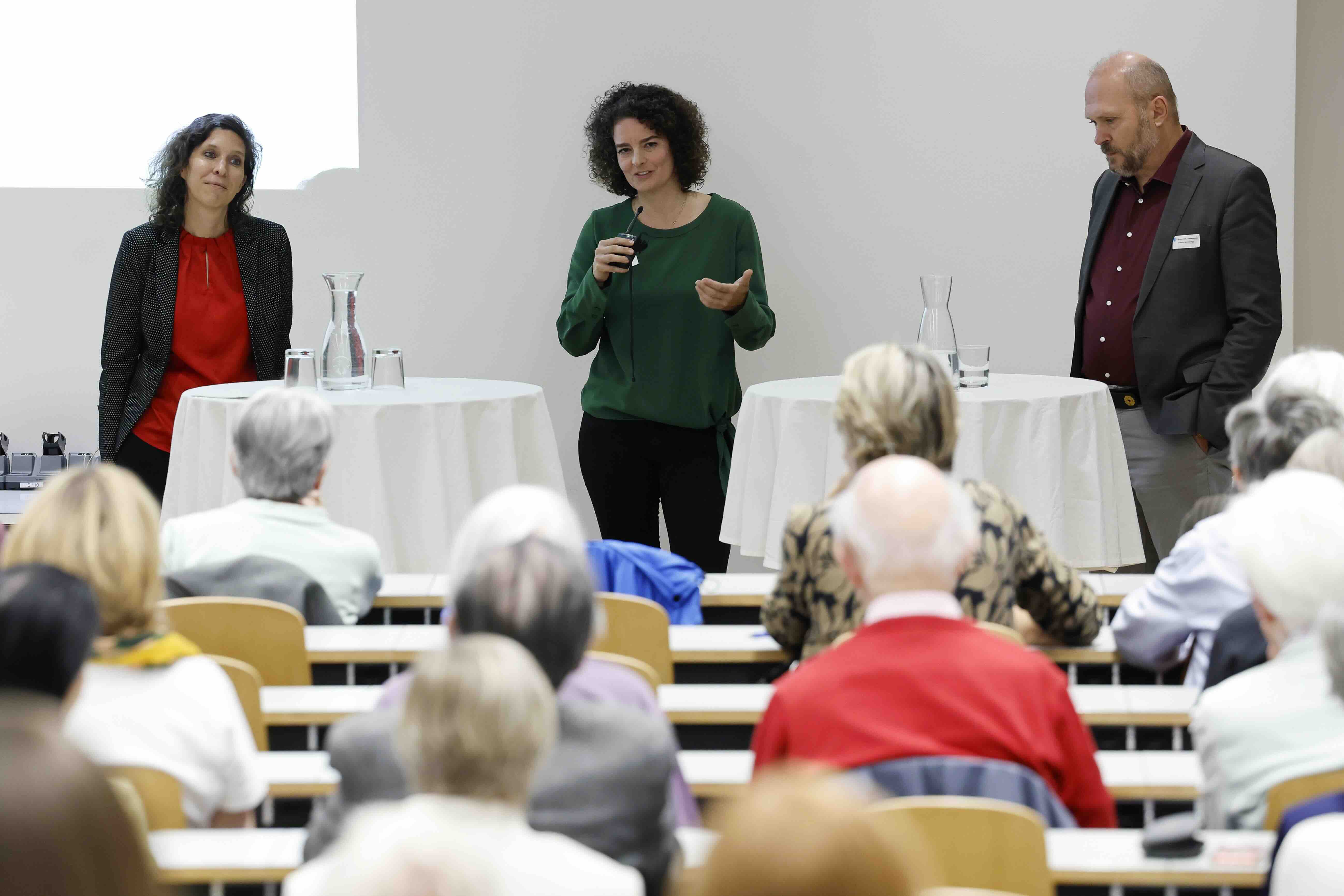 Drei Personen diskutieren bei einer Podiumsveranstaltung vor Publikum im Hörsaal.