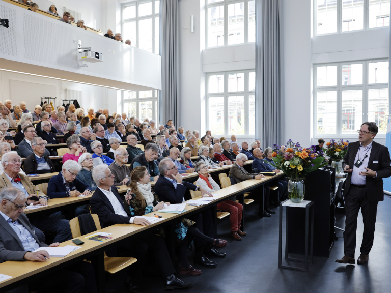 Ein Referent steht vorne in einem hellen Hörsaal und hält einen Vortrag vor einem großen, überwiegend älteren Publikum. Die Zuhörenden sitzen an langen Tischreihen und verfolgen aufmerksam die Präsentation. Der Raum ist lichtdurchflutet, mit hohen Fenstern und moderner Ausstattung. Auf einem Pult neben dem Referenten steht ein Blumenarrangement.