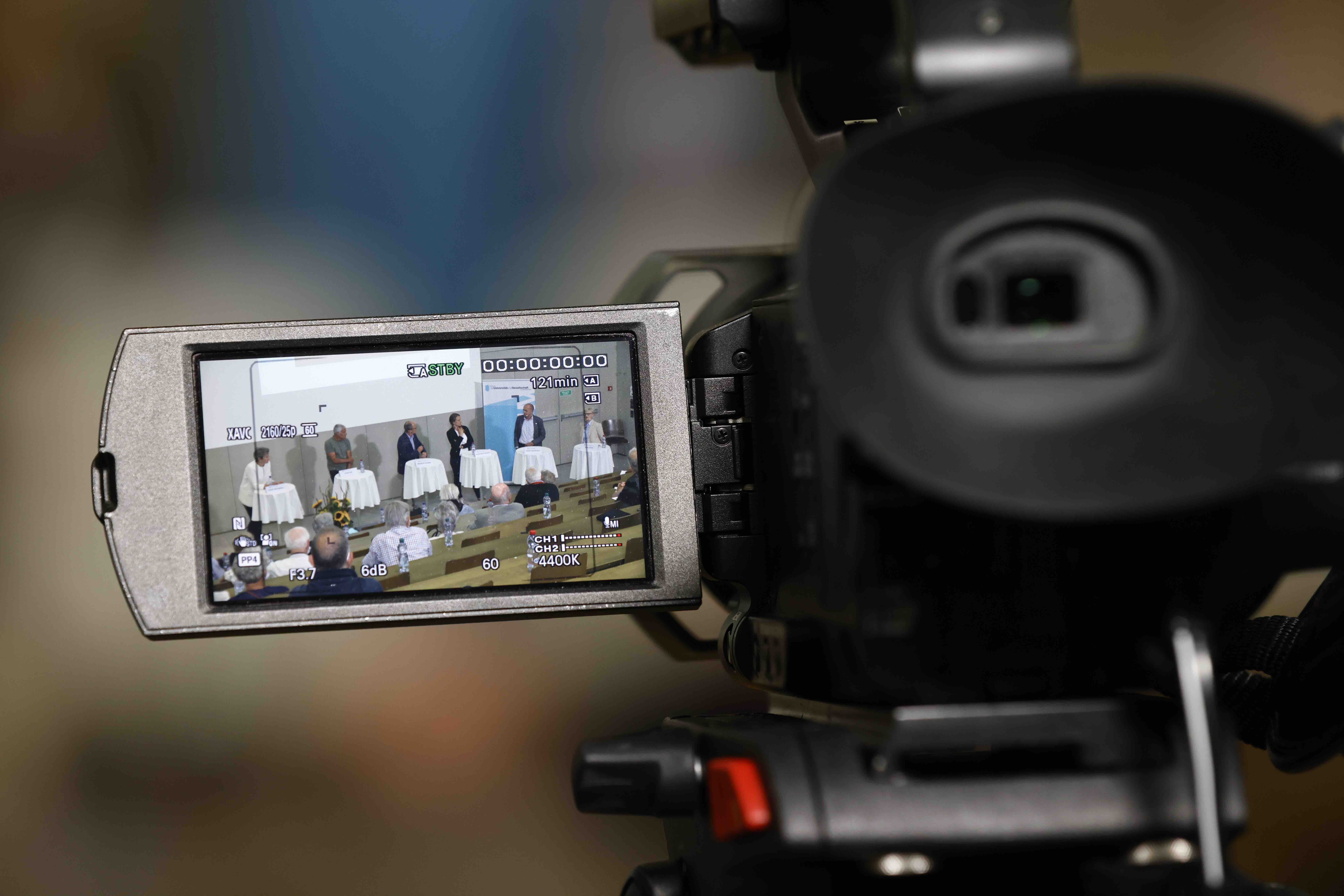 Ein Kamera-Display zeigt die Aufnahme einer Podiumsdiskussion: In einem Hörsaal stehen 6 Menschen an Stehtischen und diskutieren gemeinsam.