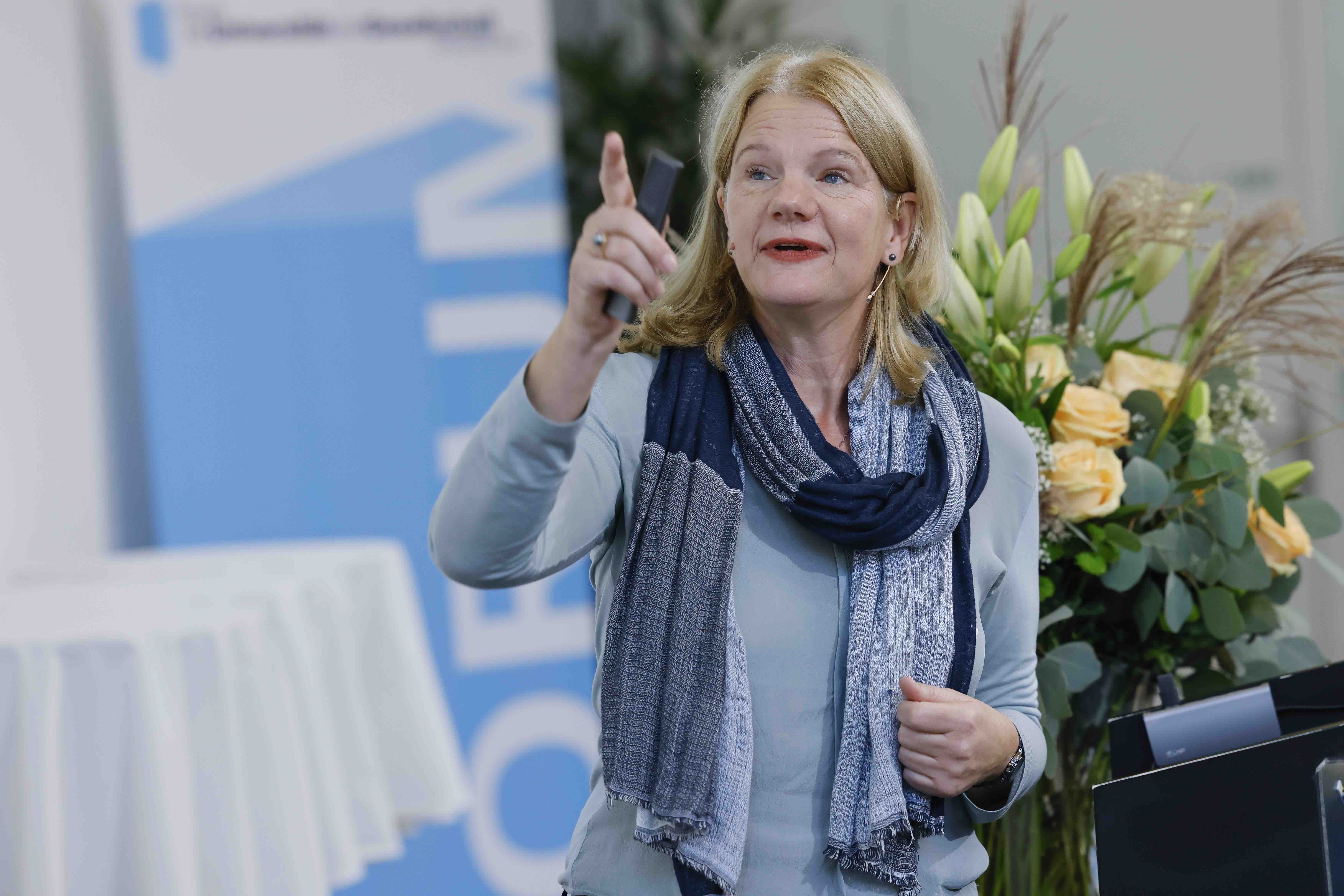 Aufnahme der Referentin Prof. Dr. Sabine Rohrmann. Sie ist blau gekleidet und gestikuliert mit der rechten Hand. Im Hintergrund sind ein Stehtisch mit weissem Tischtuch, ein Banner mit dem blauweissen Forumslogo und ein Blumenstrauss sichtbar.