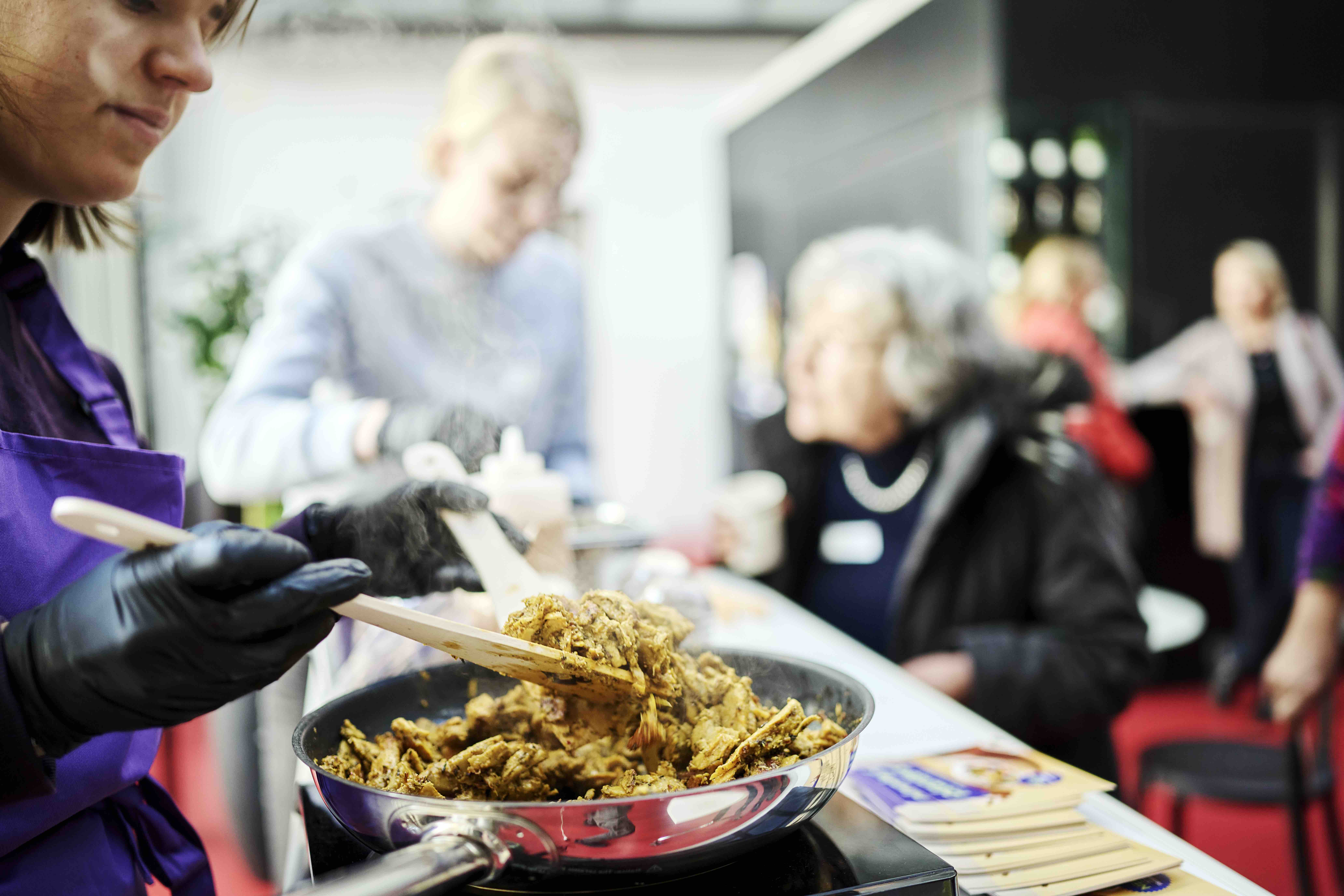 Auf einem Degustationsstand steht eine Bratpfanne mit Planted Poulet aus Erbsenprotein. Eine Frau rührt mit zwei Holzkellen, um den Stand herum stehen Menschen und schauen zu.