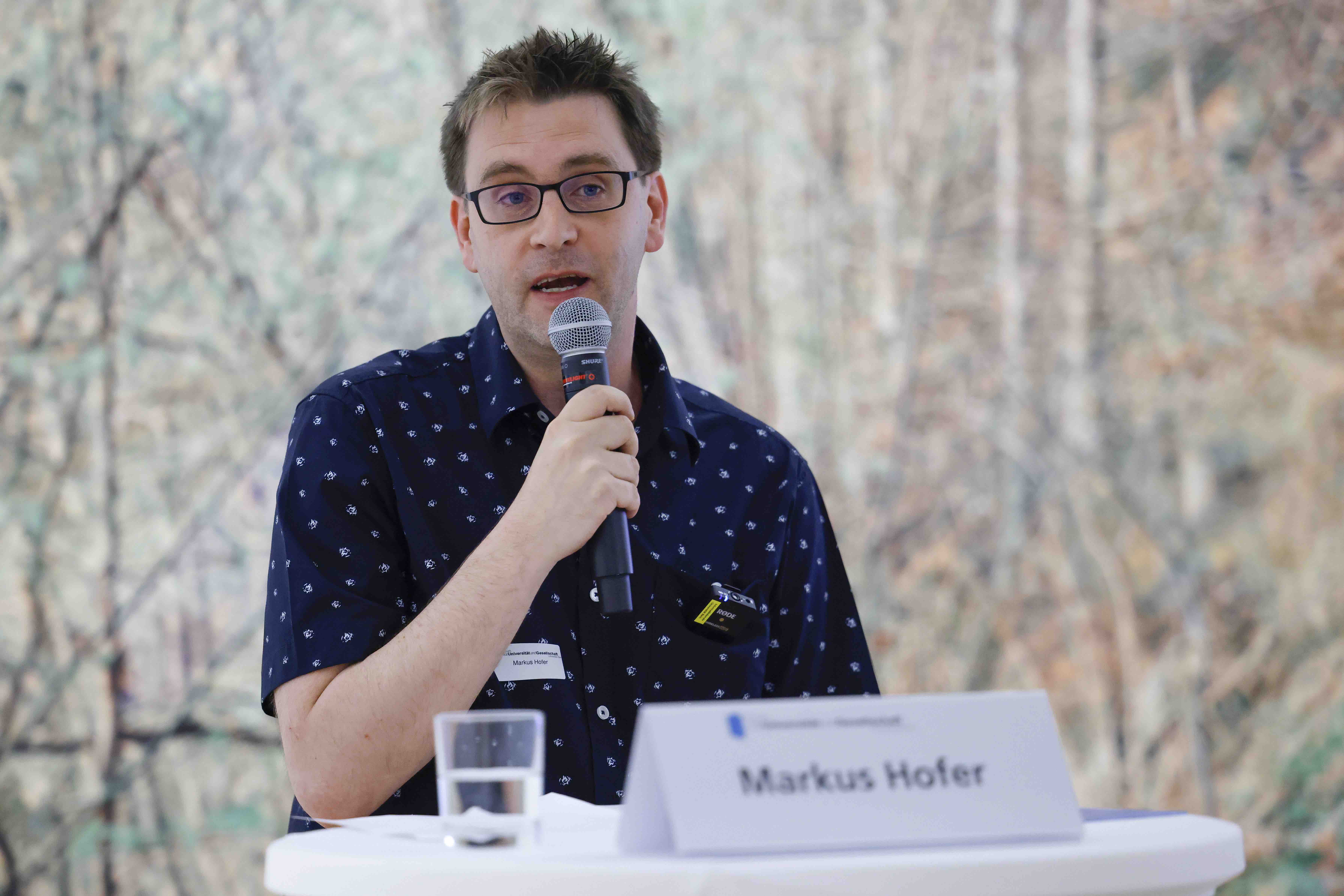 Der Referent Markus Hofer steht hinter einem Stehtisch mit weissem Tischtuch und spricht in ein Handmikrofon. Im Hintergrund ein Ausschnitt des Bildes «Frühling» von Franz Gertsch zu sehen.