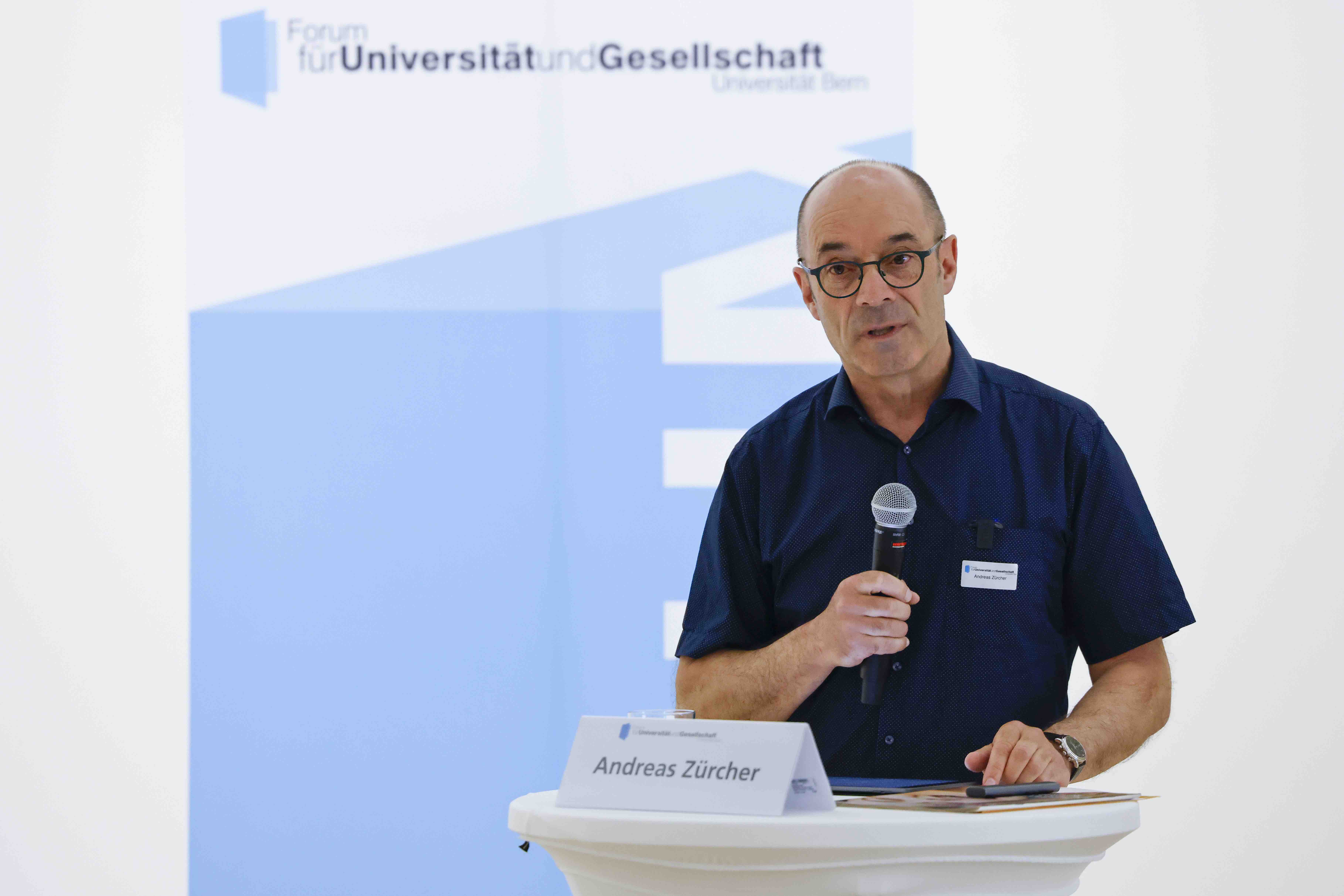 Der Referent Andreas Zürcher steht hinter einem Stehtisch mit weissem Tischtuch und spricht in ein Handmikrofon. Im Hintergrund ist das blau-weisse Logo des Forums für Universität und Gesellschaft sichtbar.