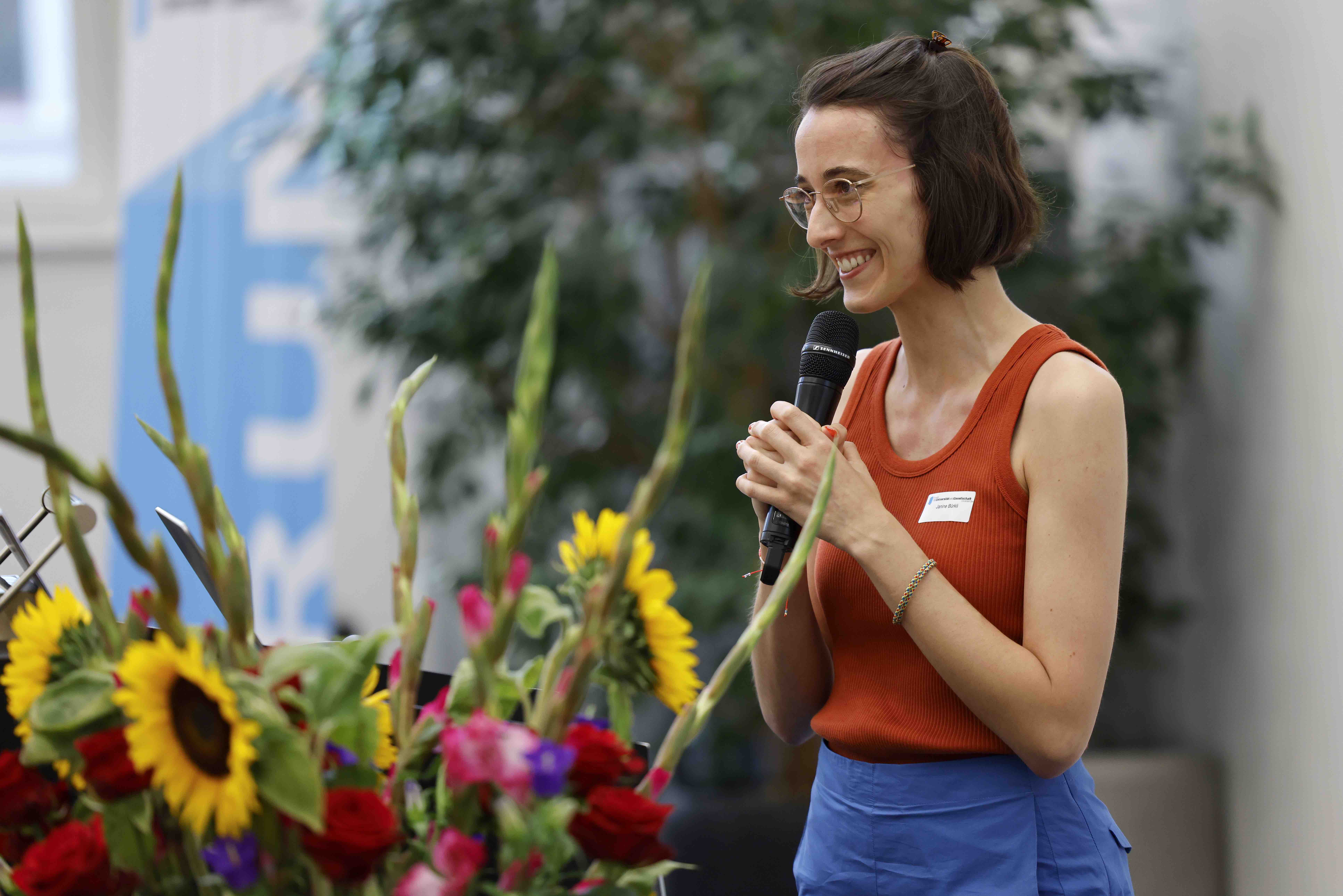 Die Referentin Janine Bürkli spricht in ein Handmikrofon und lacht. Im Hintergrund sind ein Blumenstrauss, grüne Zimmerpflanzen und ein blauweisses Banner mit dem Forumslogo erkennbar.