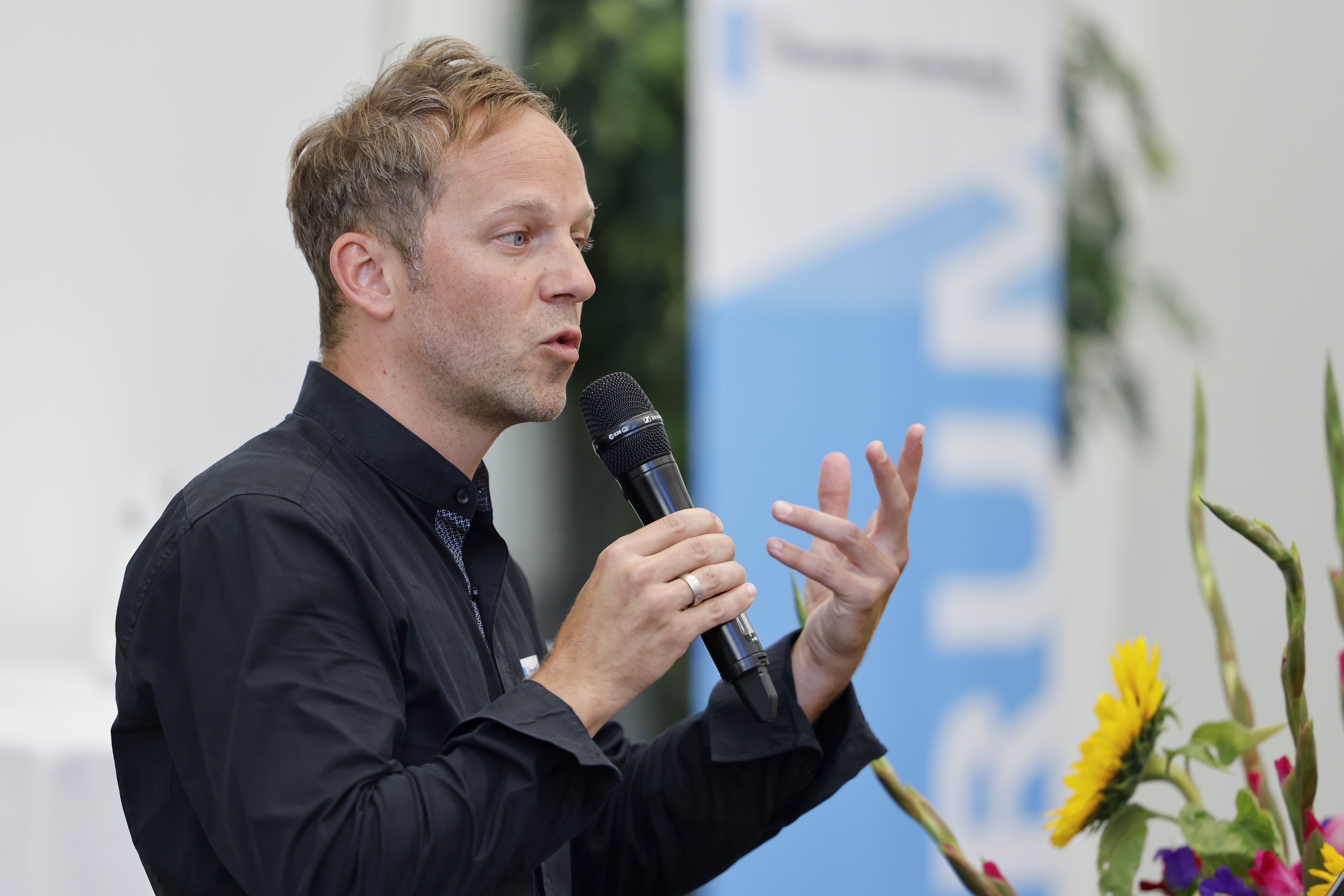 Der Referent Philipp Moll steht hinter einem Rednerpult und spricht in ein Handmikrofon. Mit der linken Hand gestikuliert er, im Hintergrund sind ein Blumenstrauss und ein Banner mit dem blauweissen Forumslogo zu erkennen.