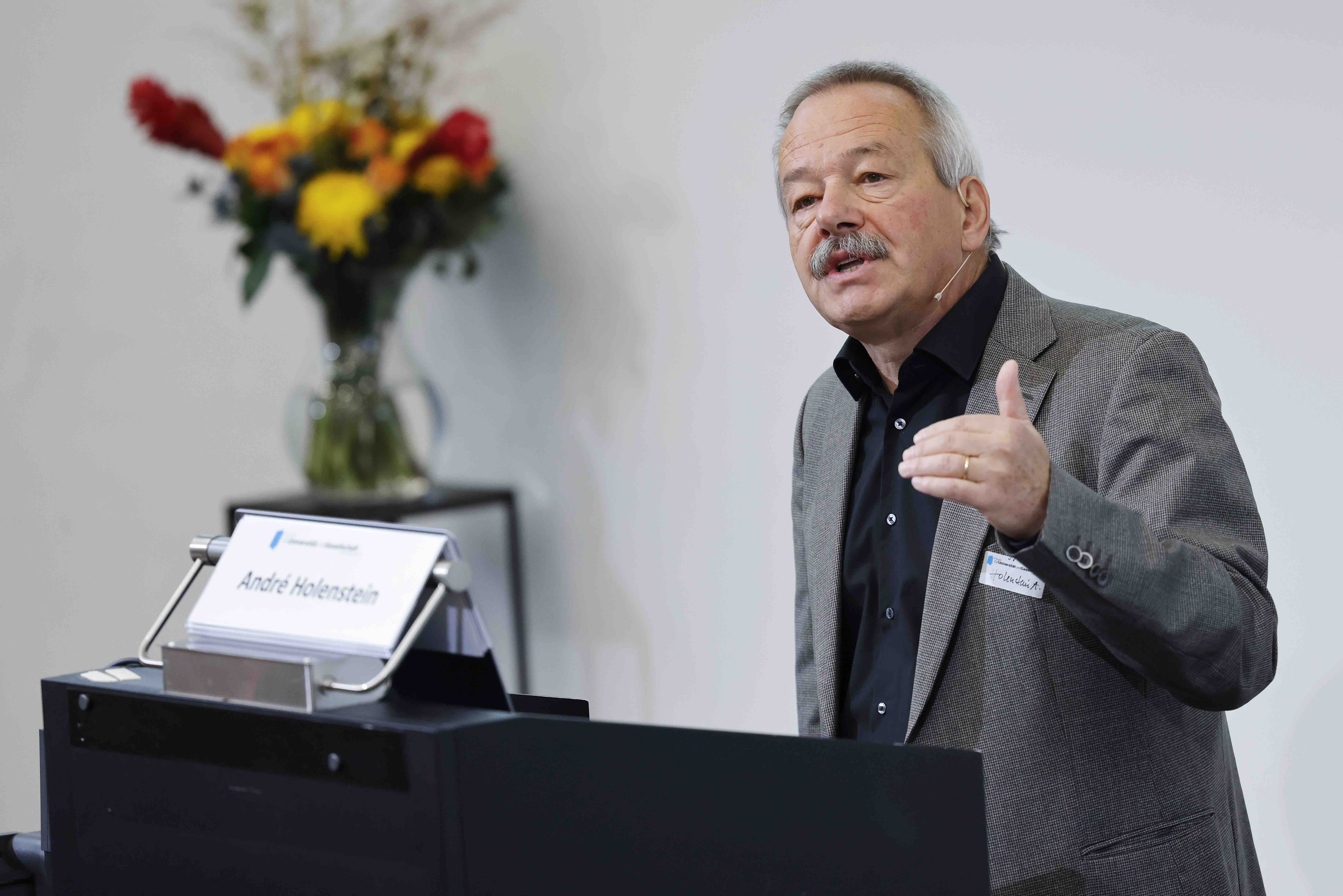 Der Referent André Holenstein steht hinter einem Rednerpult und gestikuliert mit der linken Hand. Auf der rechten Seite ist im Hintergrund ein rot-gelber Blumenstrauss
